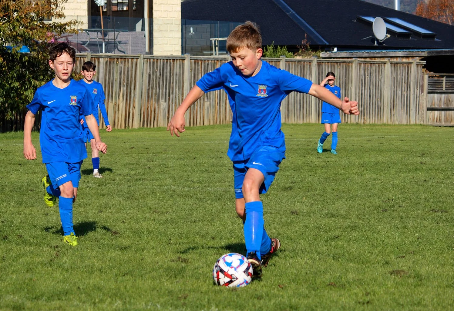Children playing soccer on a green field wearing blue uniforms. One boy in the foreground is about to kick a soccer ball, while others are running behind him. A wooden fence and houses are visible in the background.