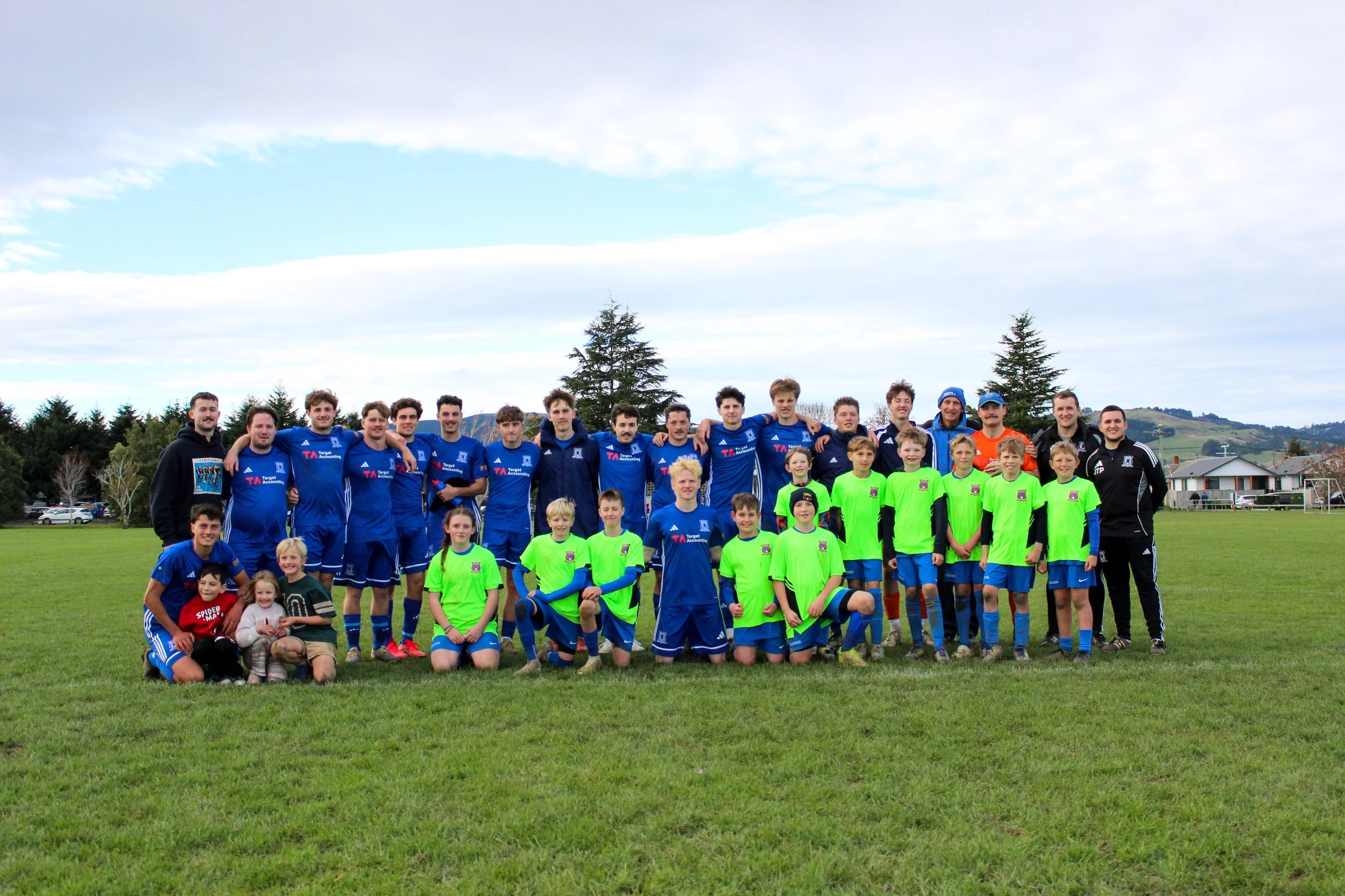 Group of soccer players and children on a field with trees and a cloudy sky in the background.