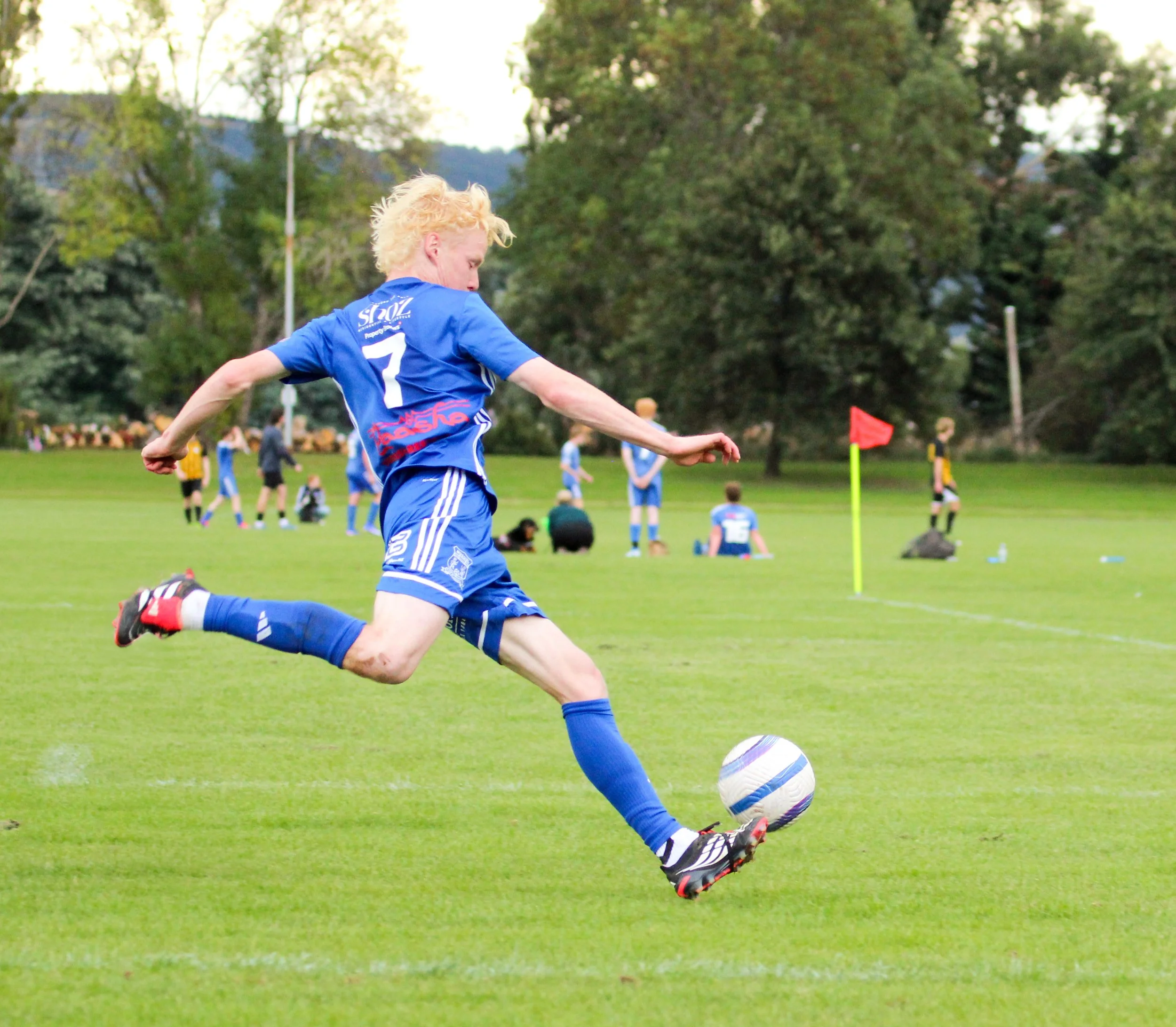 A young male soccer player with blonde curly hair wearing a blue jersey with the number 7 is kicking a soccer ball on a green field. In the background, other players and a red corner flag are visible, with trees and a partly cloudy sky.