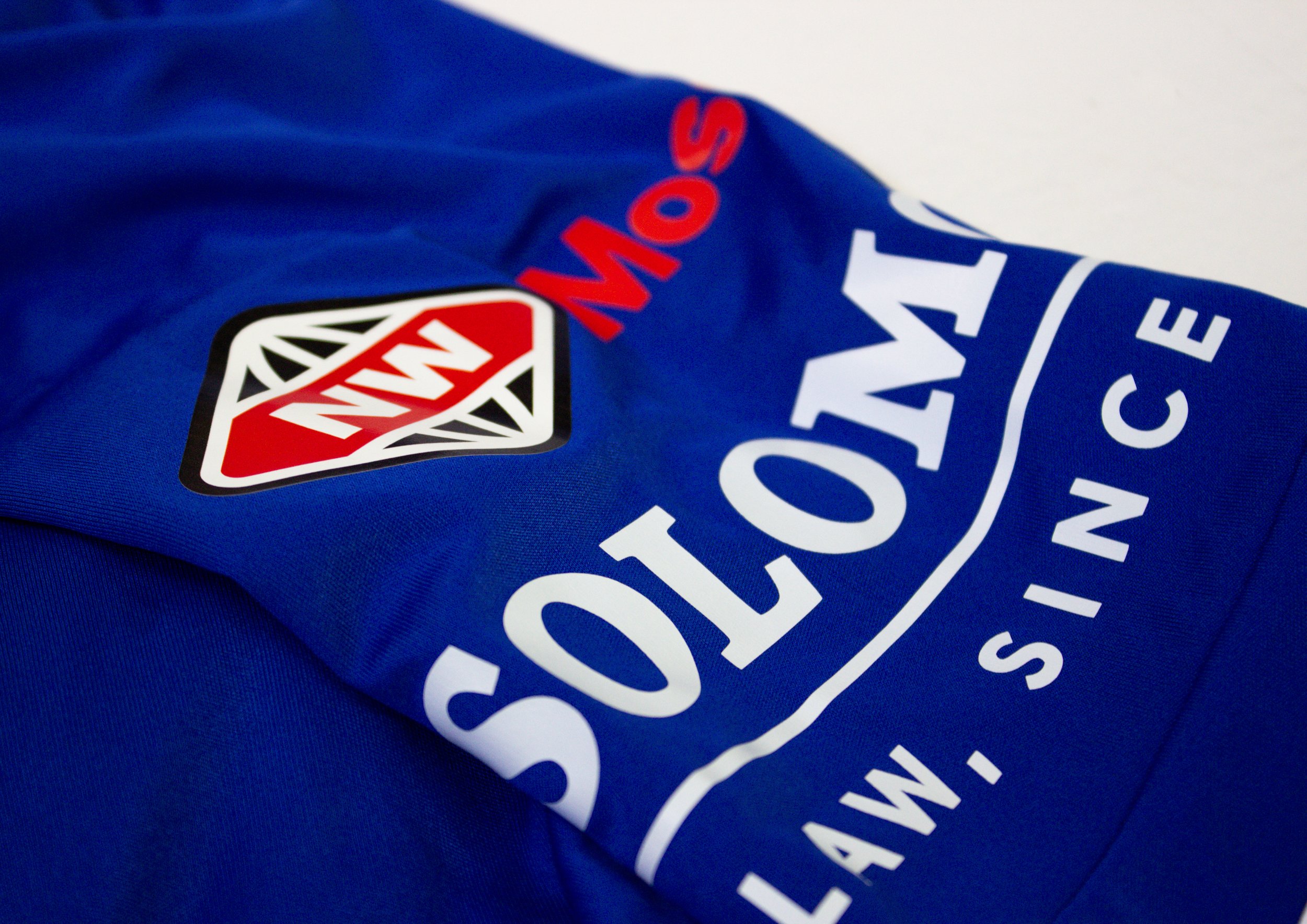 Close-up of a blue fabric with a white and red logo and text. The logo features a stylized red diamond with black and white details. The text includes the words 'STADIUM' and other partially visible words.
