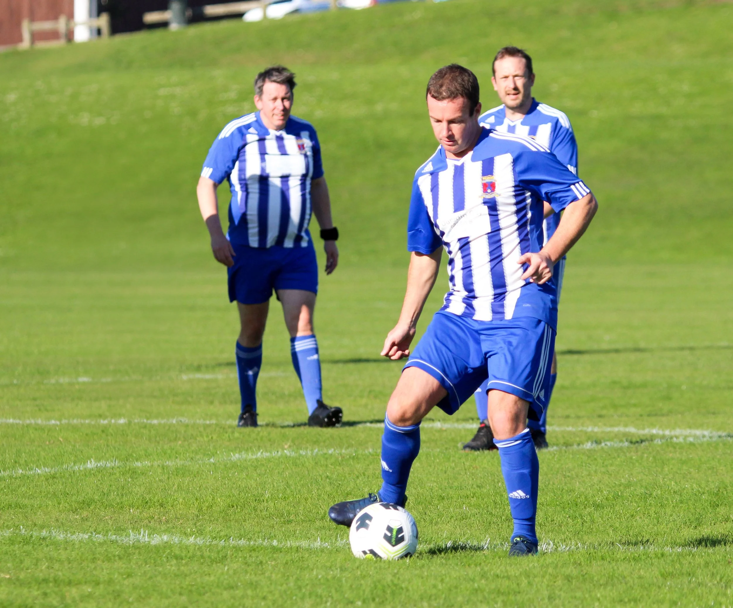 Three soccer players dressed in blue and white uniforms on a green soccer field, with one player preparing to kick a soccer ball.