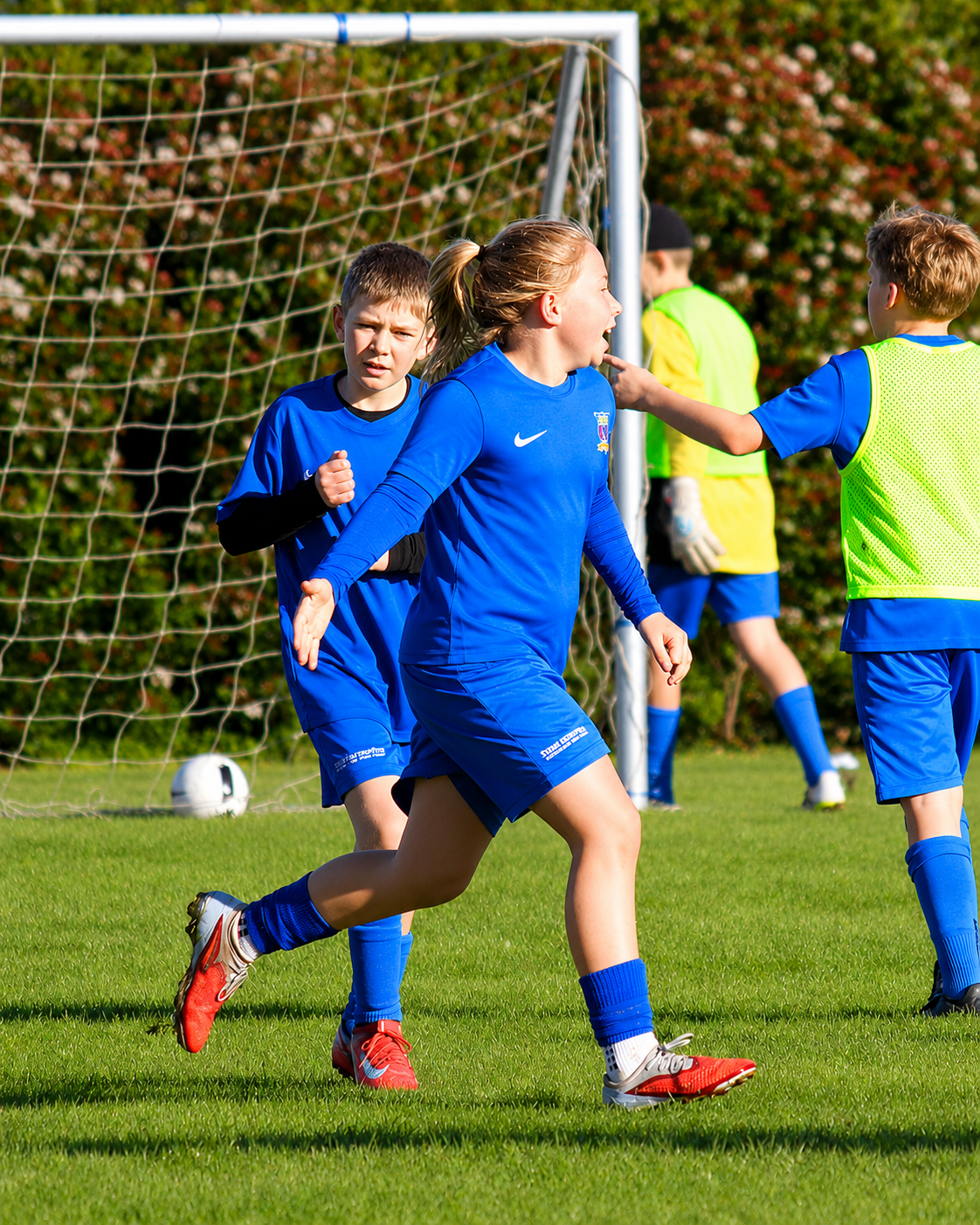 Young girls celebrating a goal in a blue soccer uniform on a grass field, with a soccer net and trees in the background.