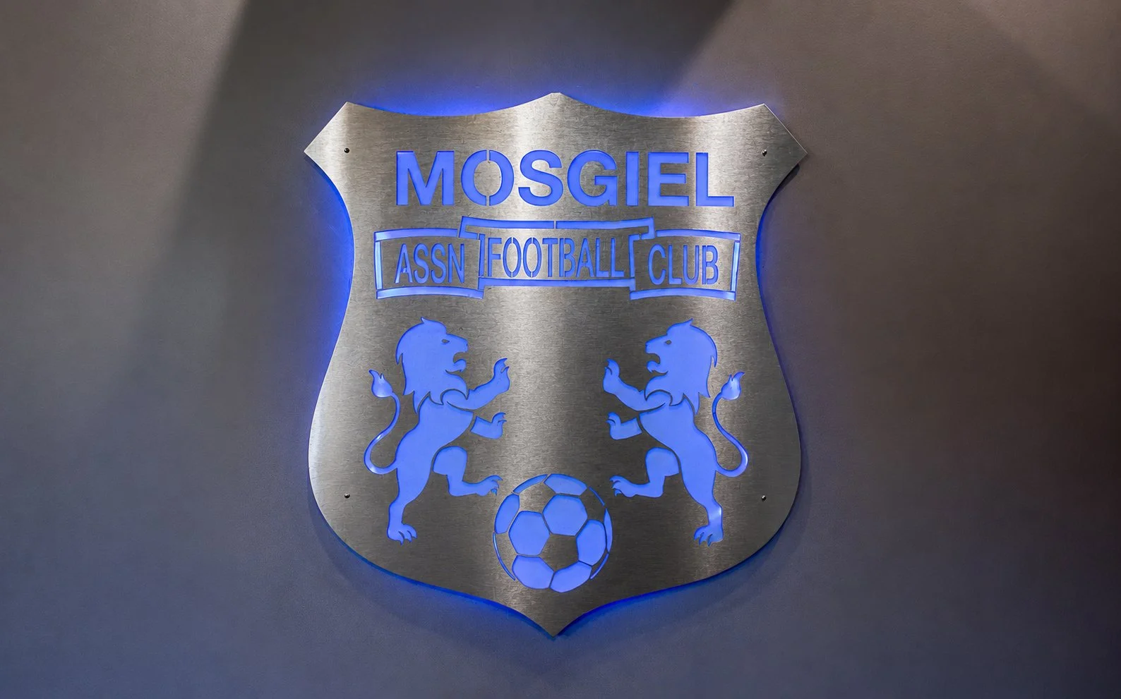 Metal emblem featuring the name 'MOSGIEL' and 'ASSN FOOTBALL CLUB' with two blue lions standing on hind legs, facing each other, holding a soccer ball at the bottom, set against a gray background with blue backlighting.