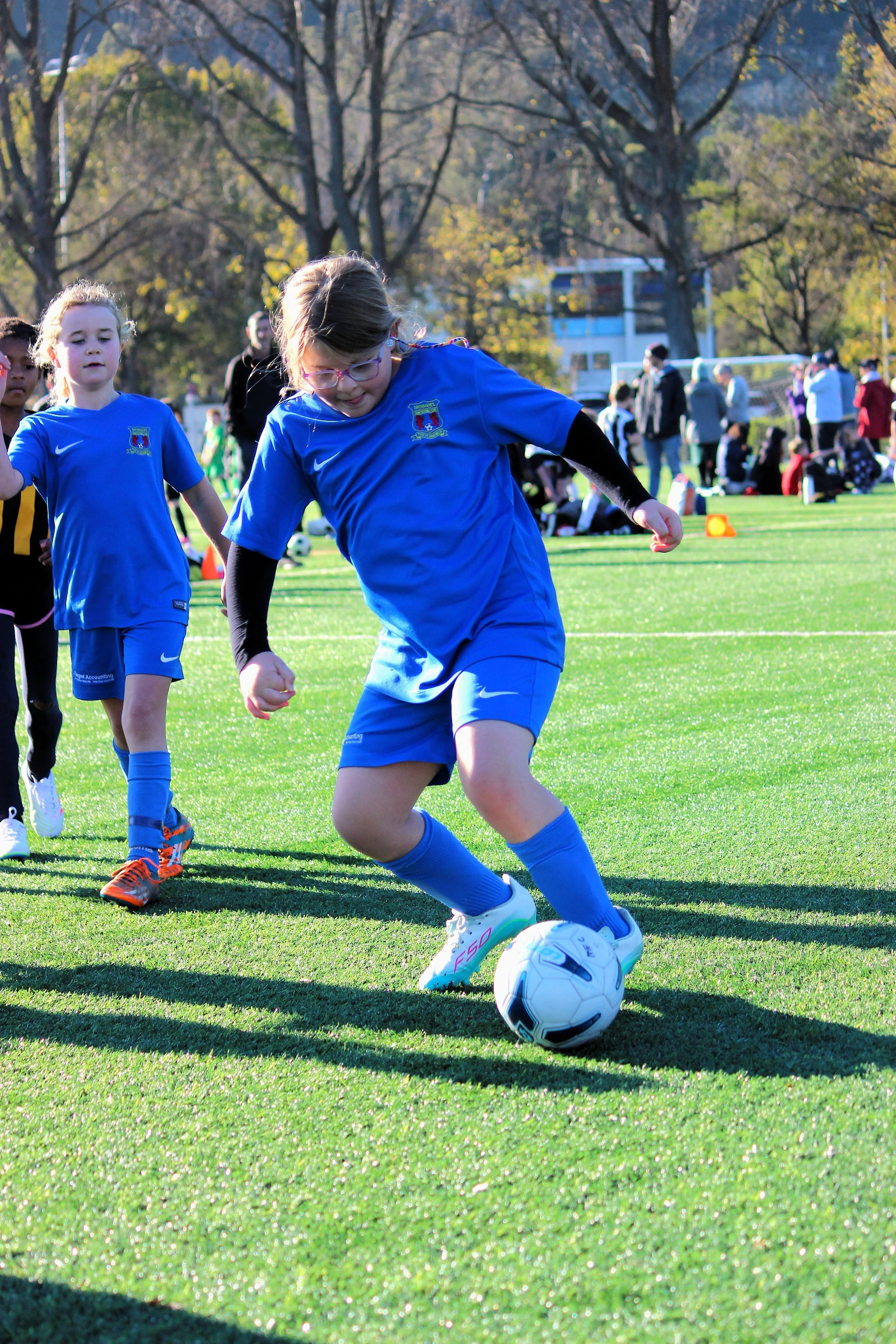 Young girl wearing glasses and a blue soccer uniform kicks a soccer ball on a grassy field during a game, with other children, parents, and trees in the background.