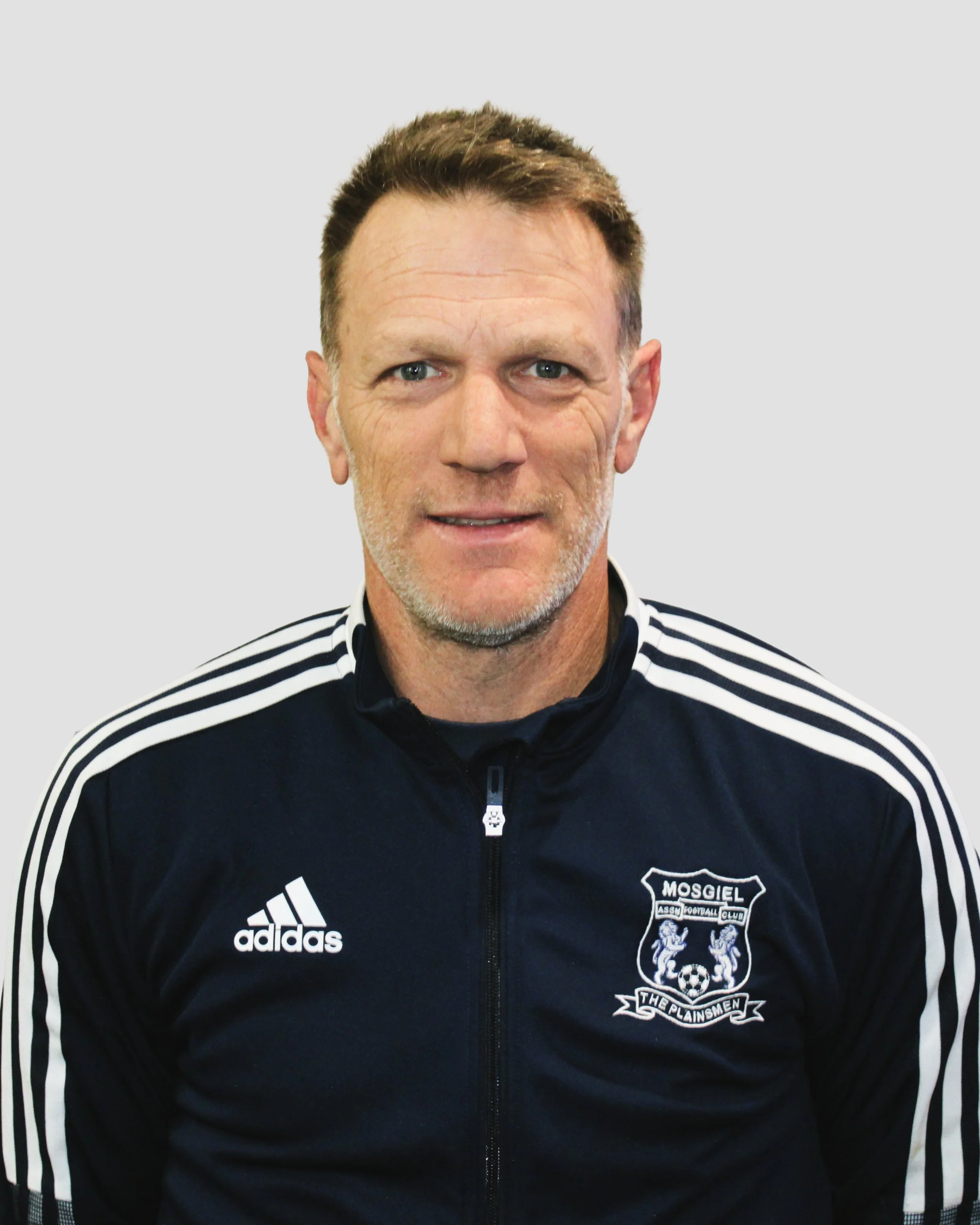 A man wearing a black Adidas sports jacket with white stripes on the sleeves and a crest of the Mosgiel Association Football Club on the chest.