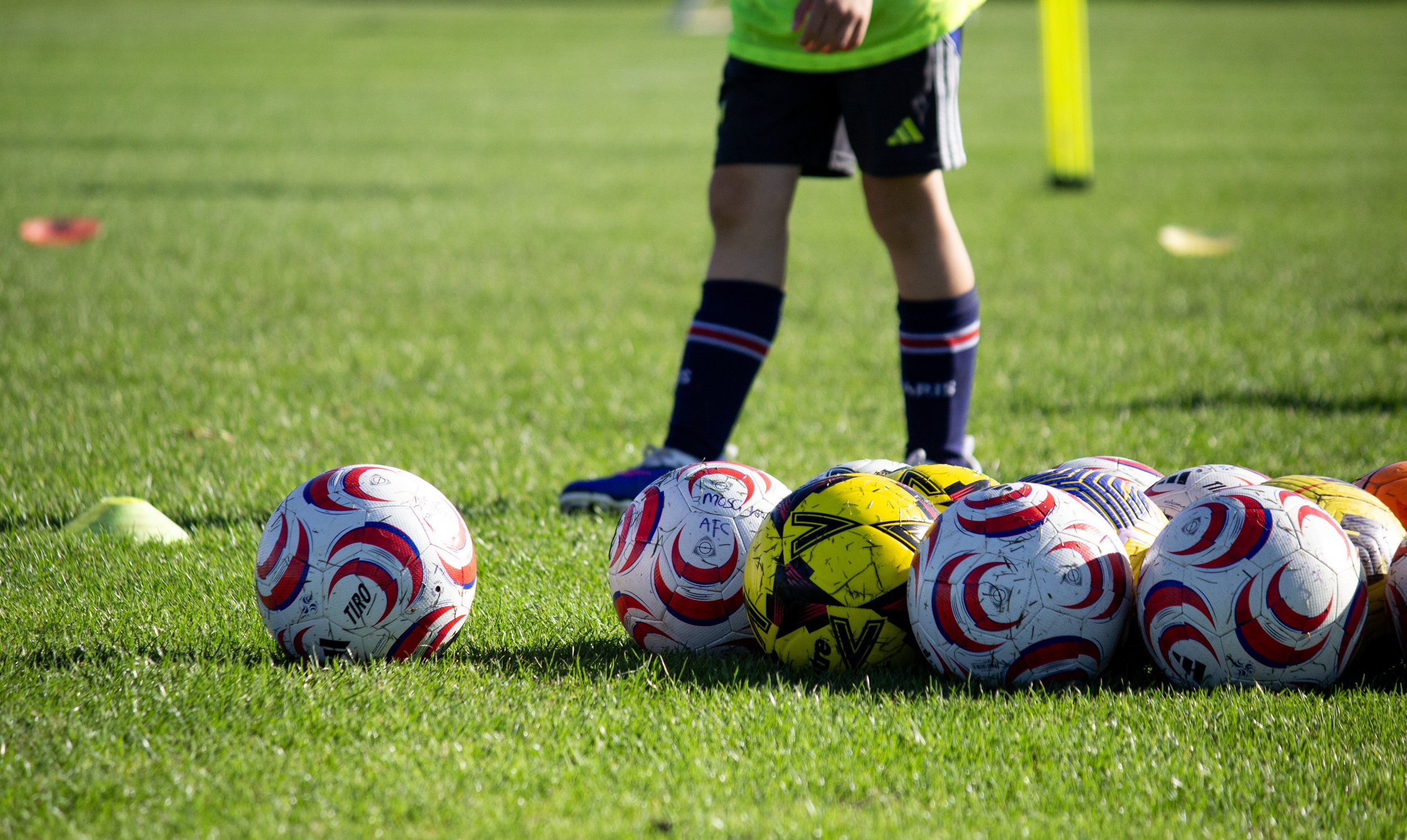 A person standing on a grassy field surrounded by multiple soccer balls, with a focus on their legs and the sports equipment.
