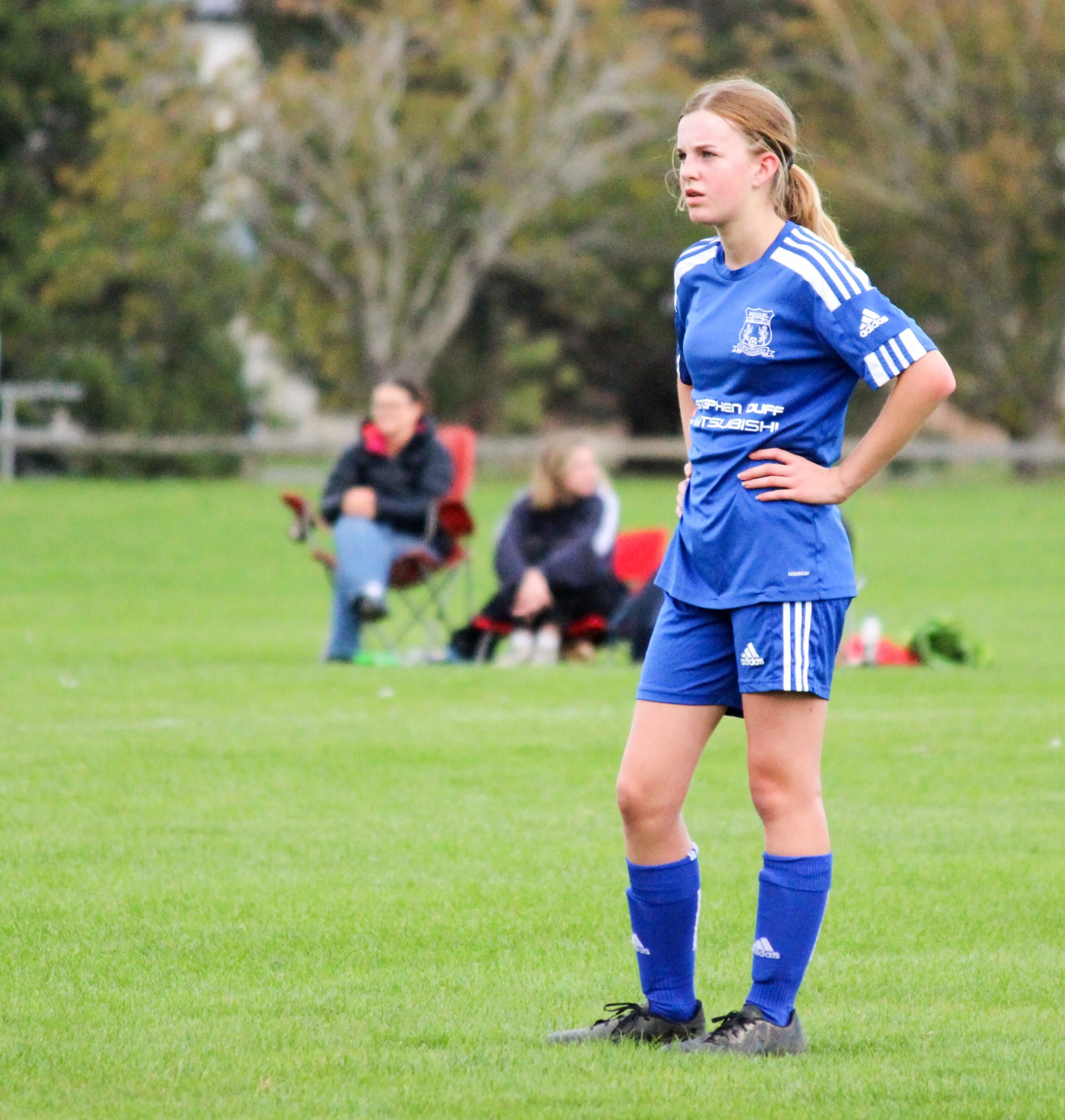 A female soccer player with red hair in a ponytail wearing a blue uniform and black cleats, standing on a grassy field with trees in the background. She appears to be focused or frustrated, with her hands on her hips.