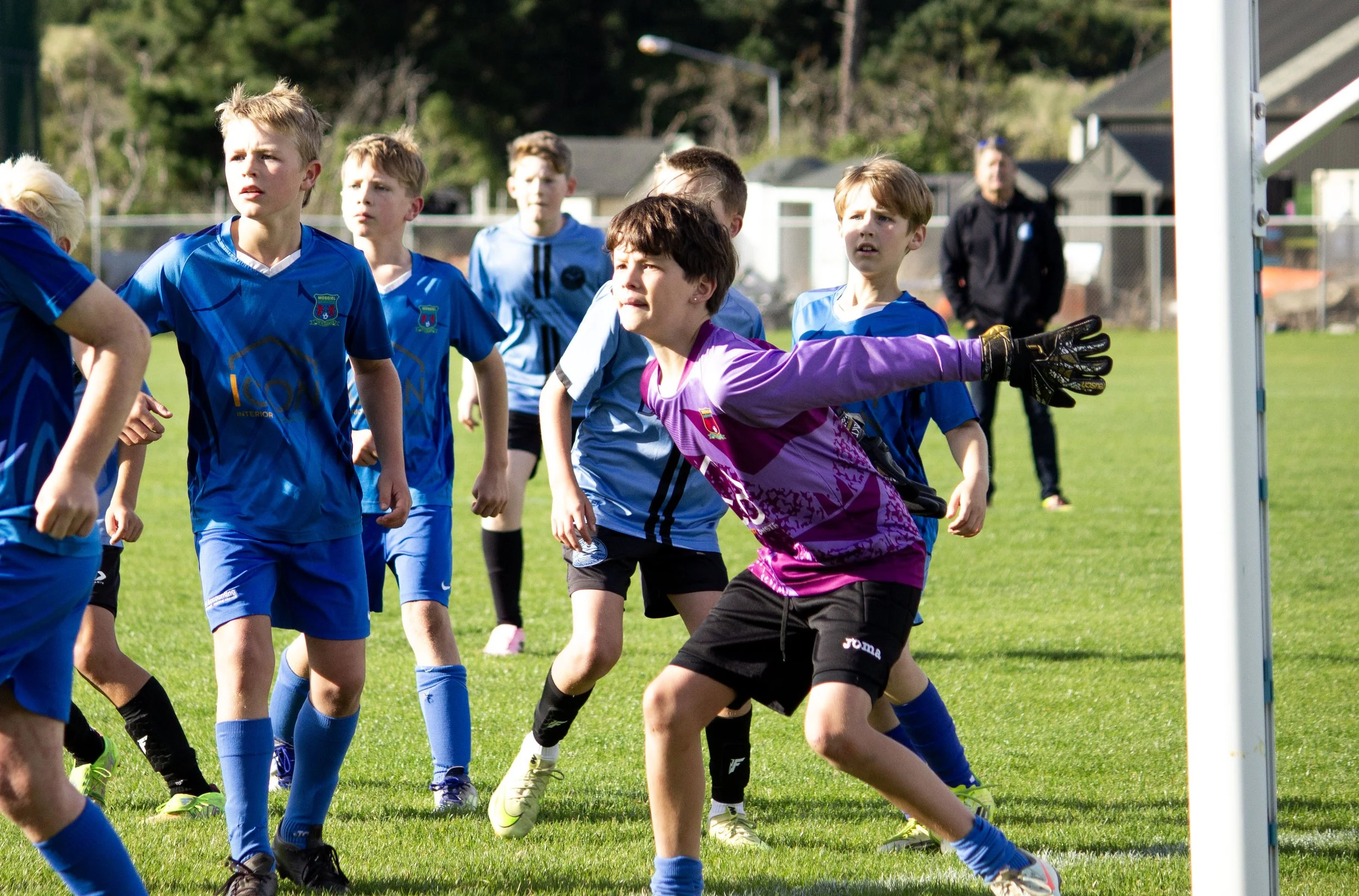 Young boys playing soccer, with a focus on a goalkeeper in purple gloves reaching out to block the ball during a game on a sunny day.