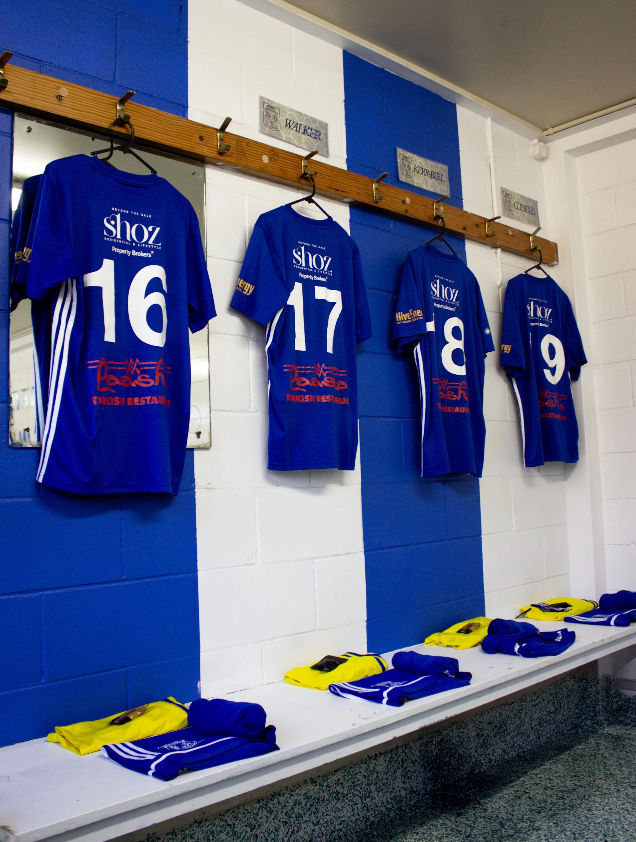 Inside a sports locker room with blue and white walls, hanging blue soccer jerseys with white numbers, and a white bench holding yellow and blue apparel.
