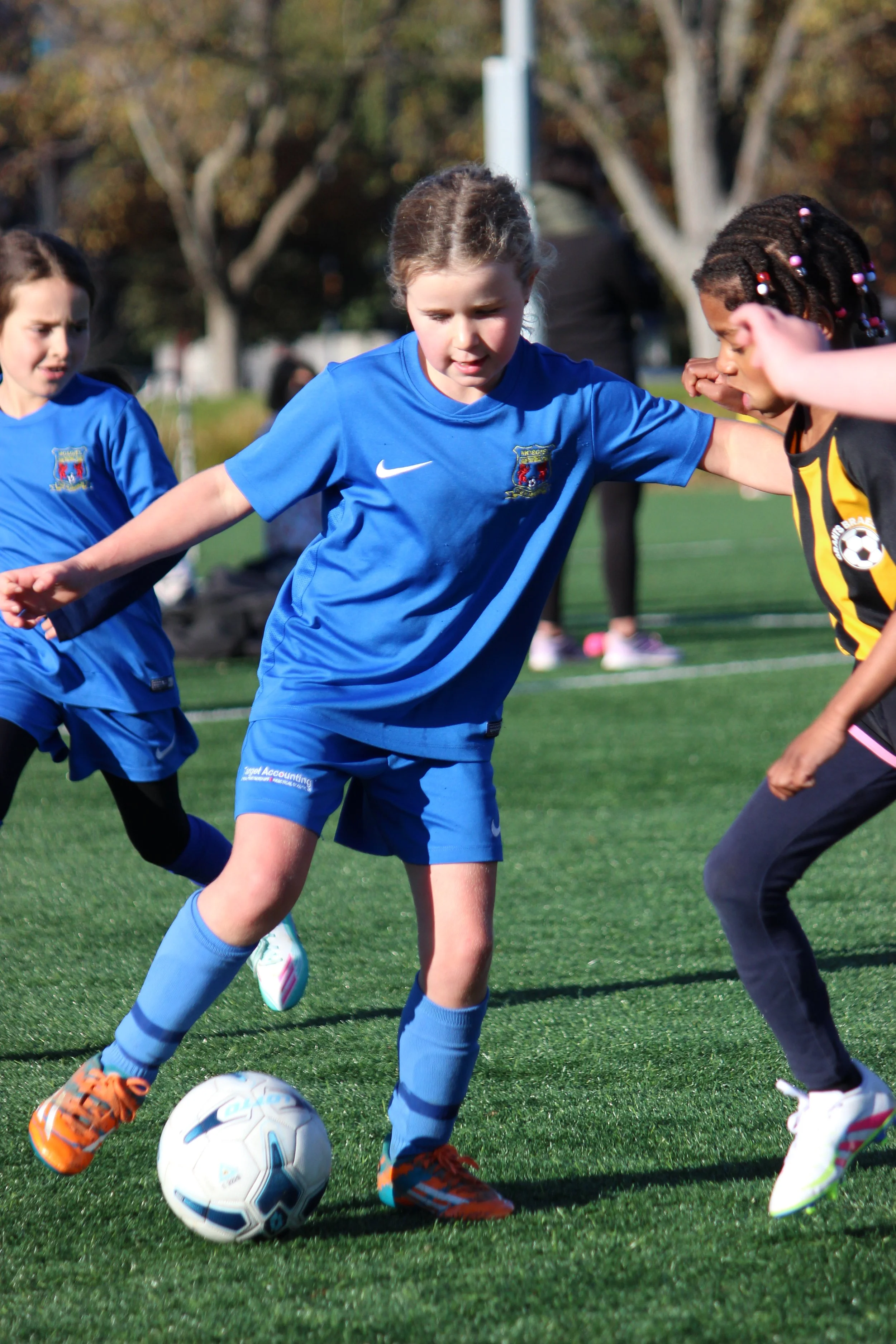 Children playing soccer outdoors, wearing sports uniforms, on a green field with trees in the background.