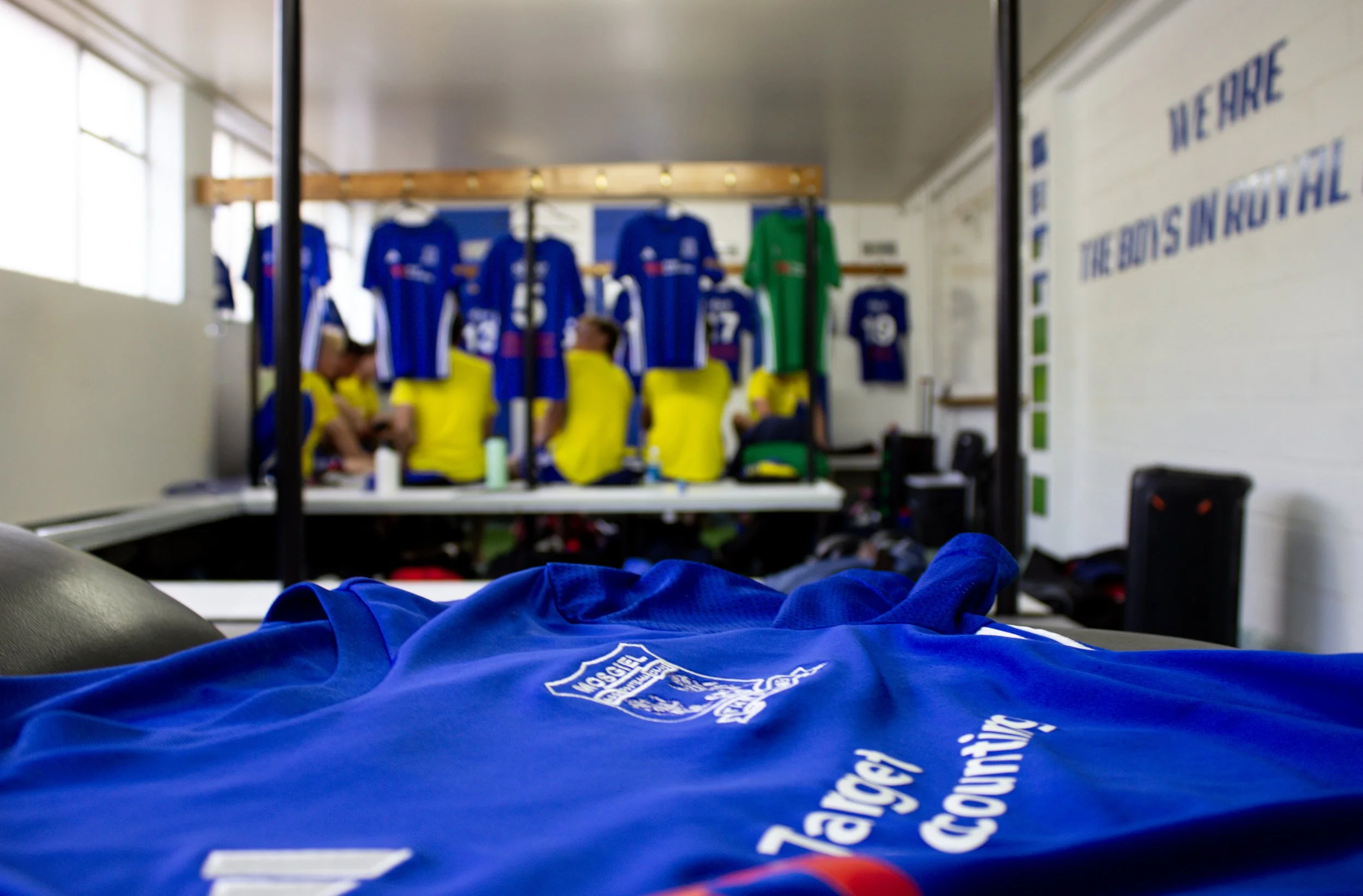 Photo shows the view from a bed or resting area in a locker room, with a blue jersey or shirt in the foreground. In the background, there are several blue and green sports jerseys hanging on a rack, with a few people seated and preparing or resting. The room has white walls, large windows, and some signage or posters.