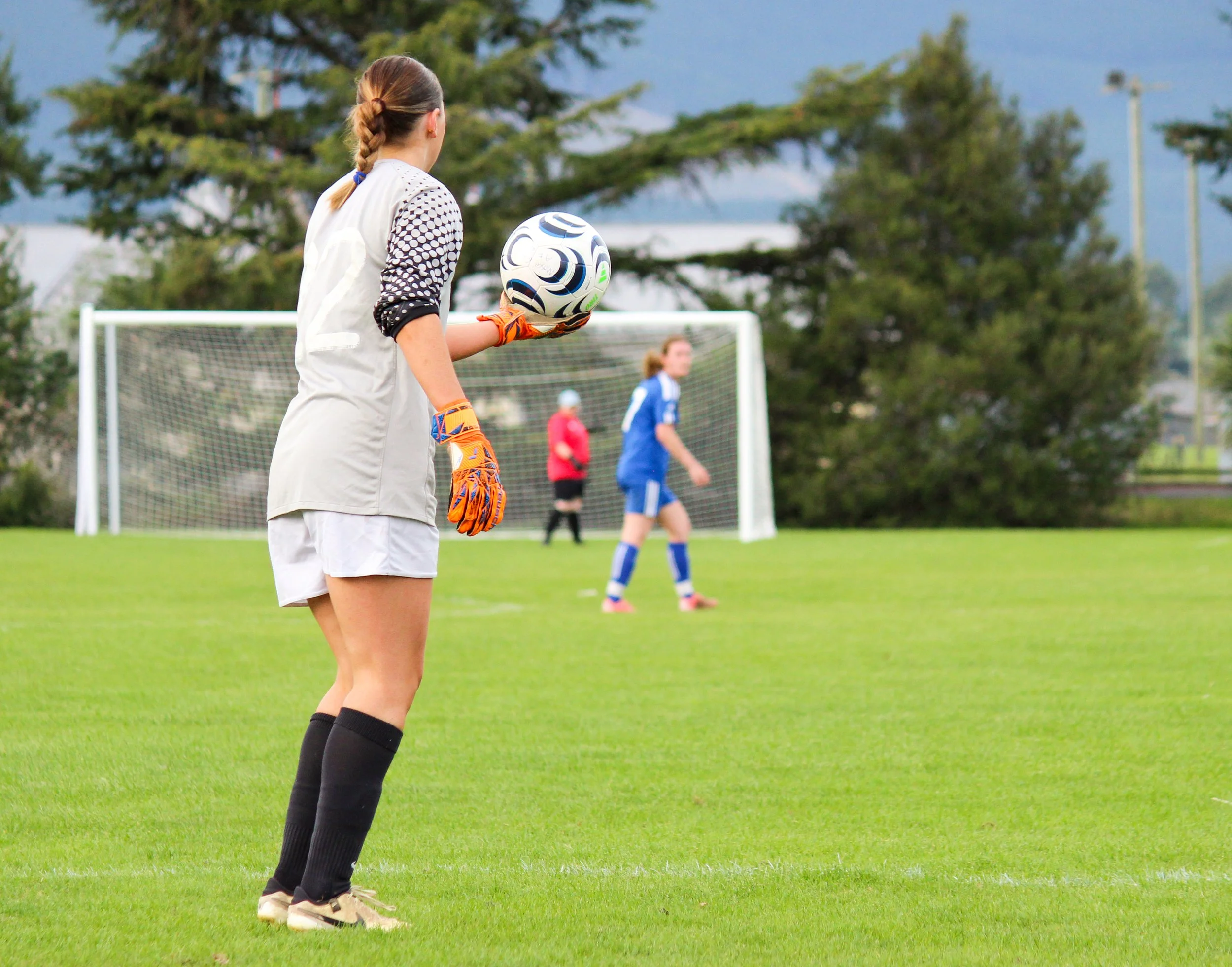 A female soccer goalkeeper standing on a soccer field holding a soccer ball, wearing gloves and a jersey, with other players and goalpost in the background.