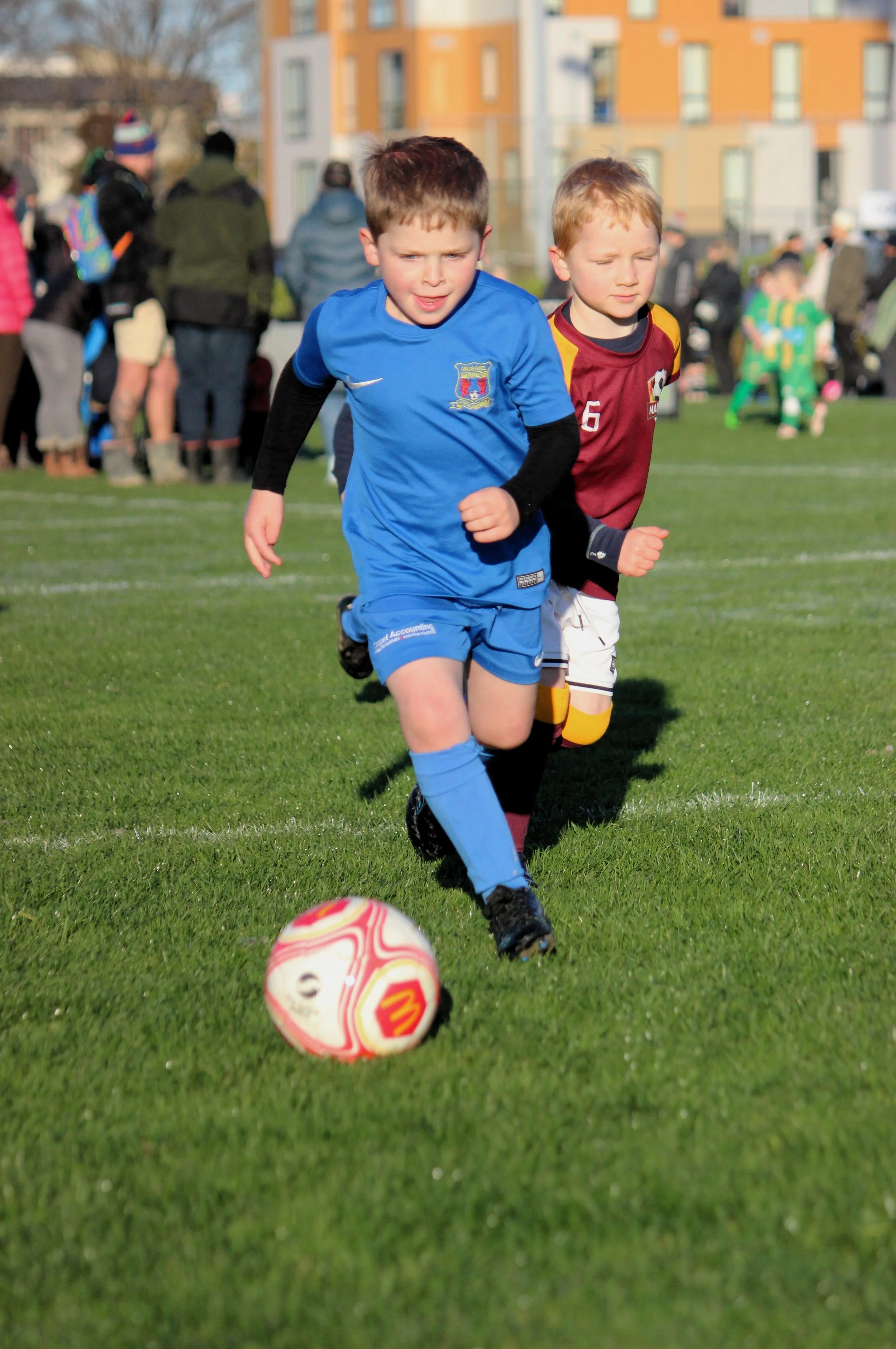 Two young boys playing soccer on a grassy field during daylight, with a ball approaching them, and a crowd of spectators and other players in the background.