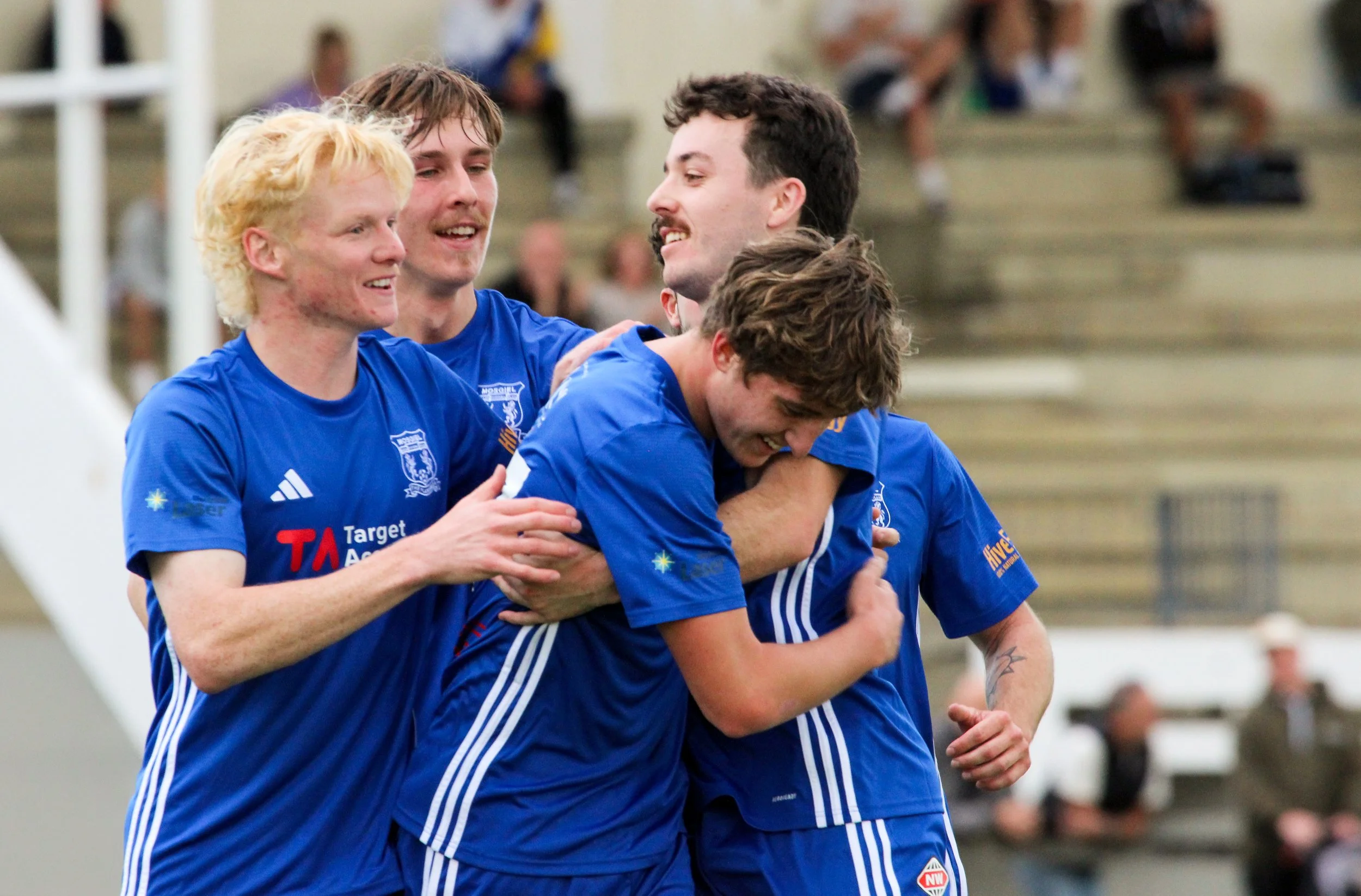 Four young men in blue sports jerseys celebrating together, hugging and smiling during a sports event in an indoor gymnasium.
