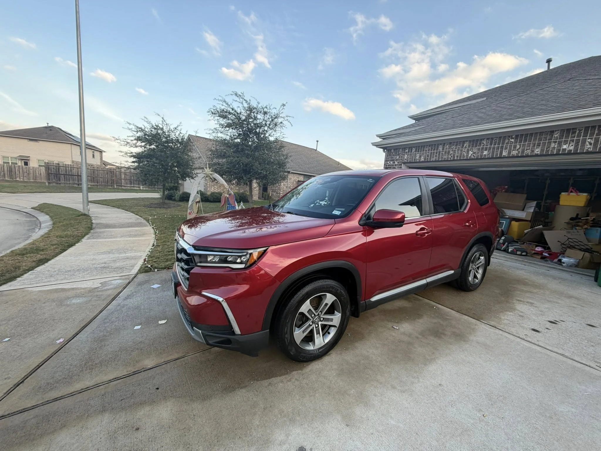 A red SUV parked in a driveway of a suburban house with an open garage filled with various items, neighboring houses, trees, a sidewalk, and a blue sky with some clouds.