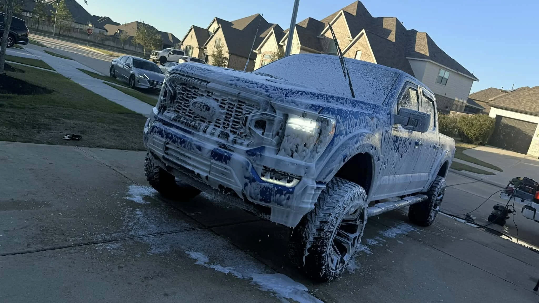 A white pickup truck covered in soap suds during a car wash in a suburban neighborhood, with houses, trees, and parked cars in the background.