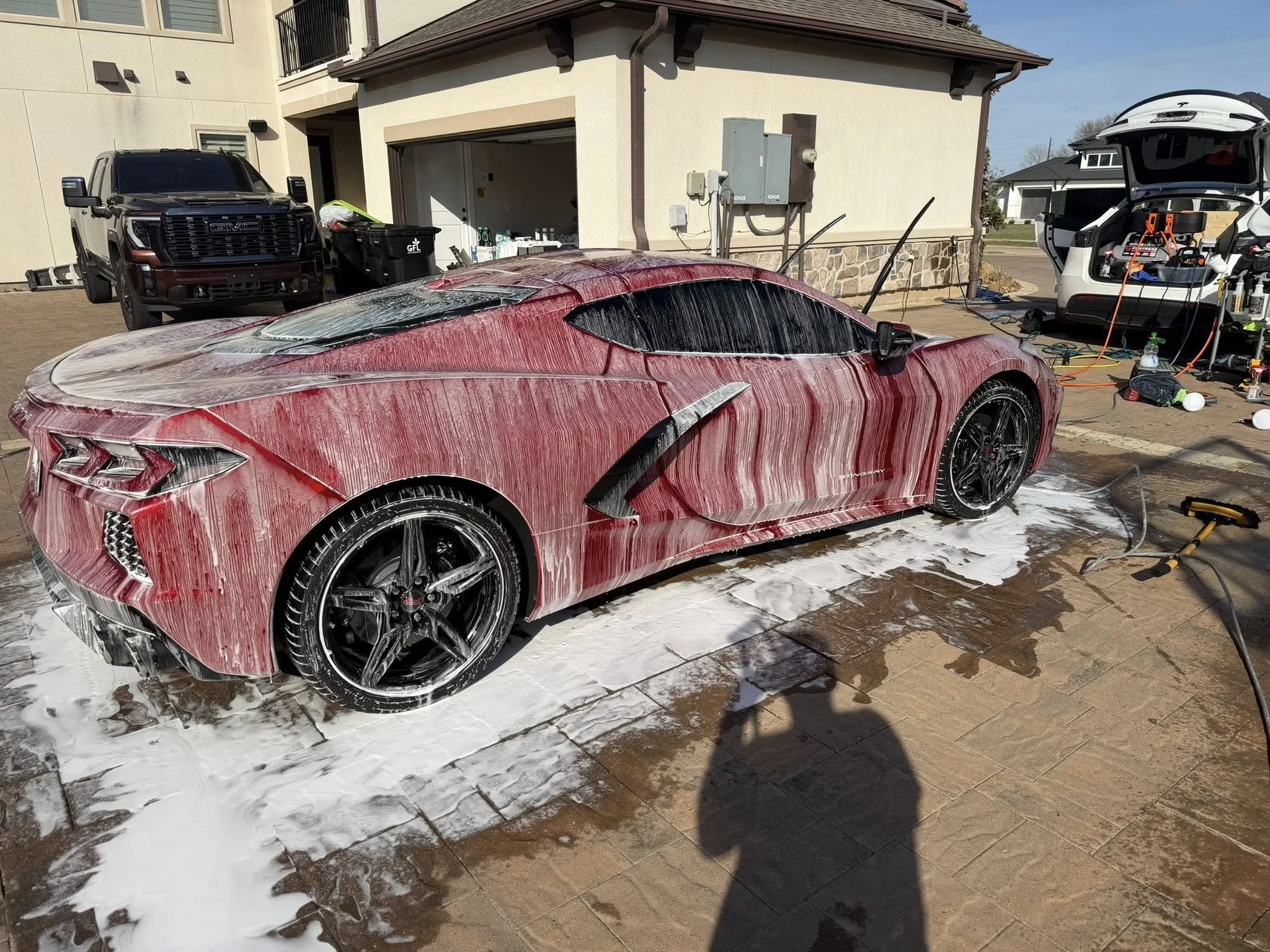 A red sports car, possibly a Lexus, is being washed with soap and water. The car is in front of a modern house with a garage and a driveway. There is a black GMC truck parked near the house and an open car trunk with cleaning equipment nearby.