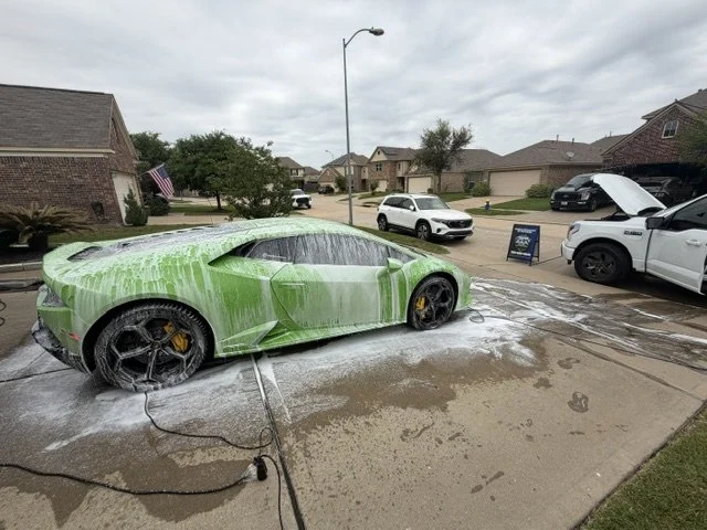 Green sports car being washed with foam and water on a driveway in a suburban neighborhood.