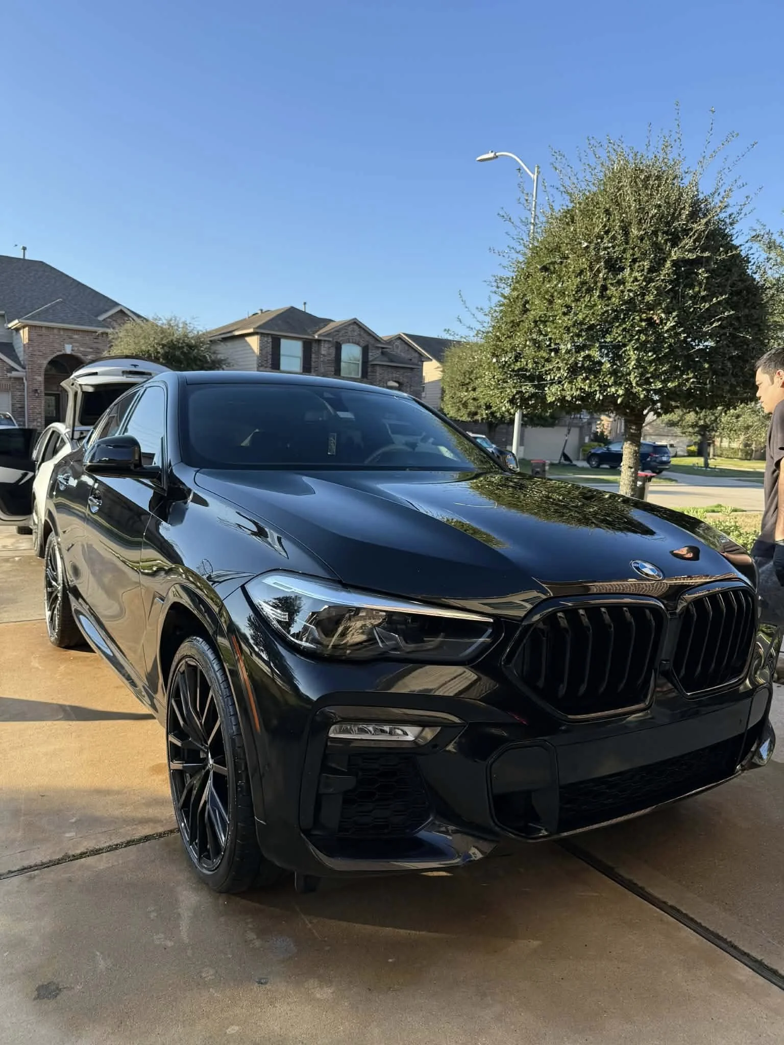 A black BMW car parked in a driveway in a suburban neighborhood with houses, trees, and a person standing nearby.
