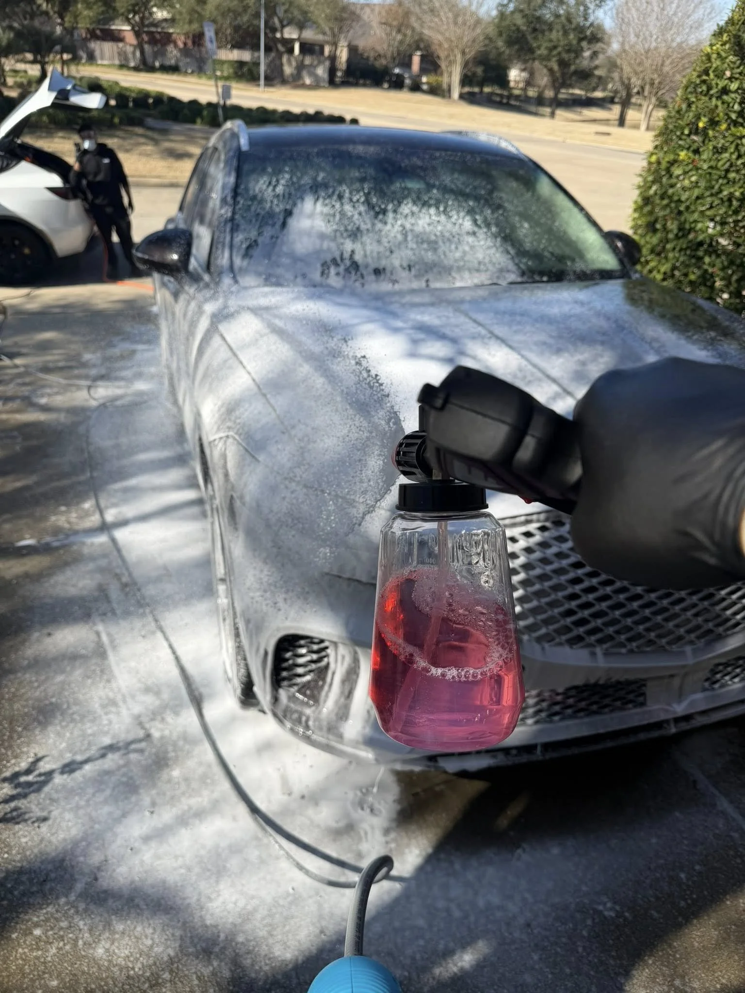 A person is using a foam cannon to wash a silver car outdoors on a sunny day. The foam cannon is attached to a hose and is dispensing pink soap foam onto the car's surface. In the background, a person is standing near another car with the trunk open, wearing a face mask, with trees and a grassy area visible.