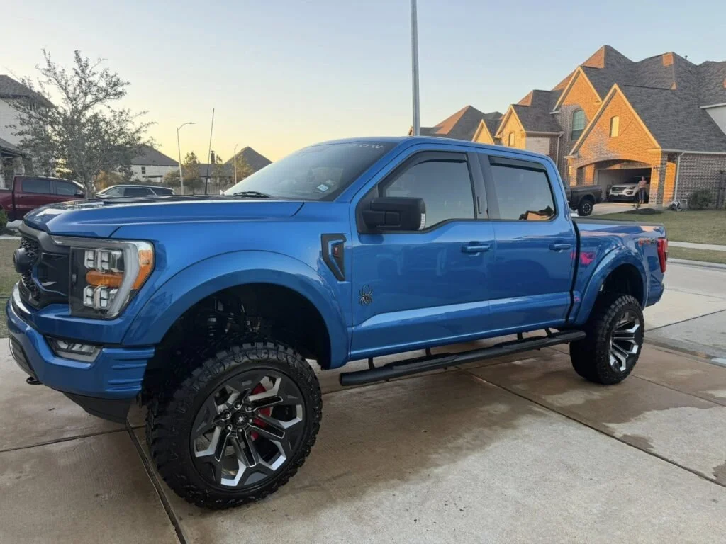 Blue lifted pickup truck with large off-road tires parked in a driveway in front of suburban houses.