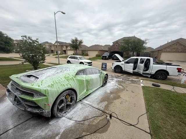 A green sports car being washed with soap and water on a driveway in a suburban neighborhood, with other cars and houses in the background.