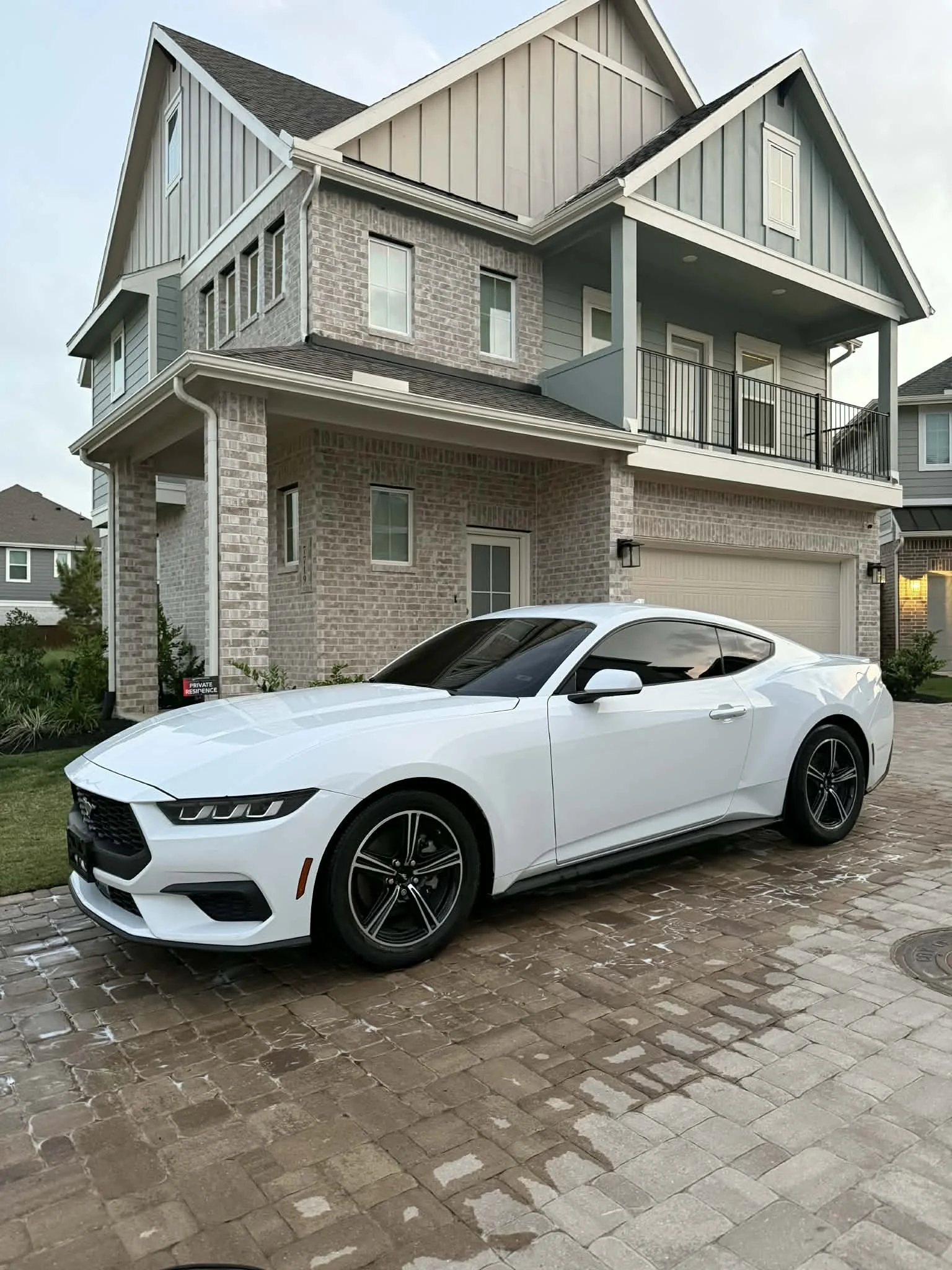 White sports car parked on a brick driveway in front of a modern two-story house with brick and gray siding exterior.
