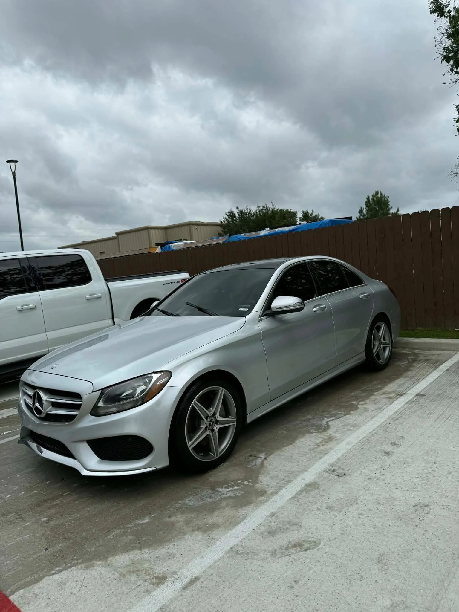 A parking lot with a silver Mercedes-Benz car in the foreground, a white truck, and a wooden fence in the background. Overcast sky with gray clouds.