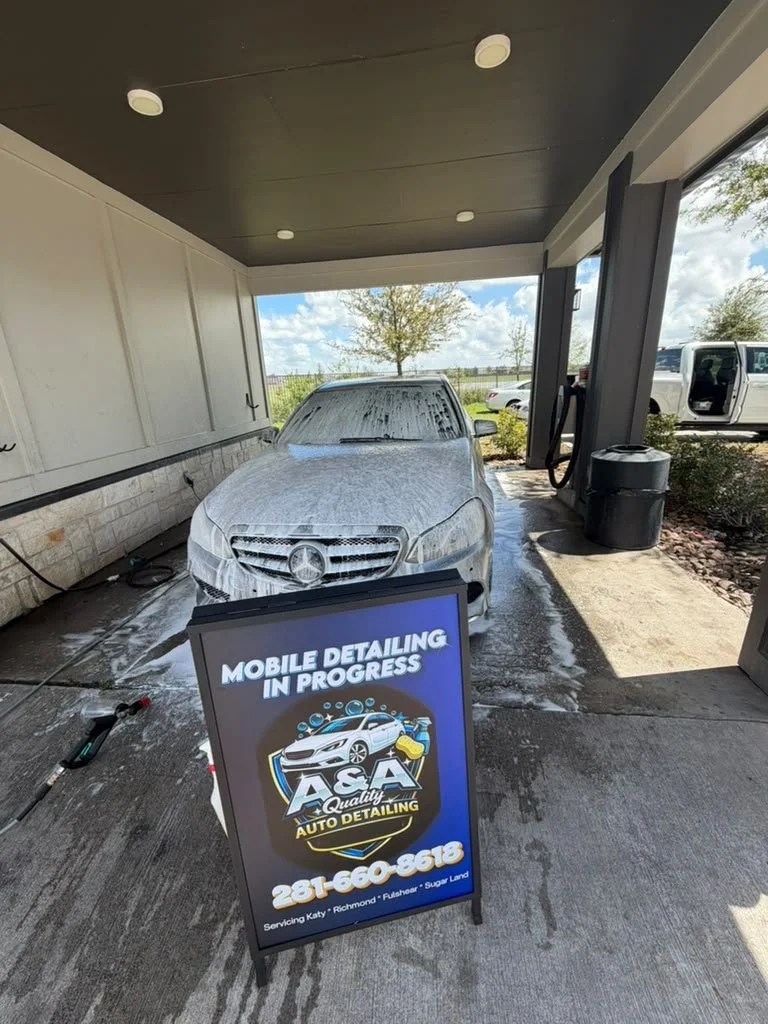 Car wash station with a silver Mercedes-Benz covered in soap suds, under a covered outdoor area, with a sign advertising A&A Quality Auto Detailing and a phone number.