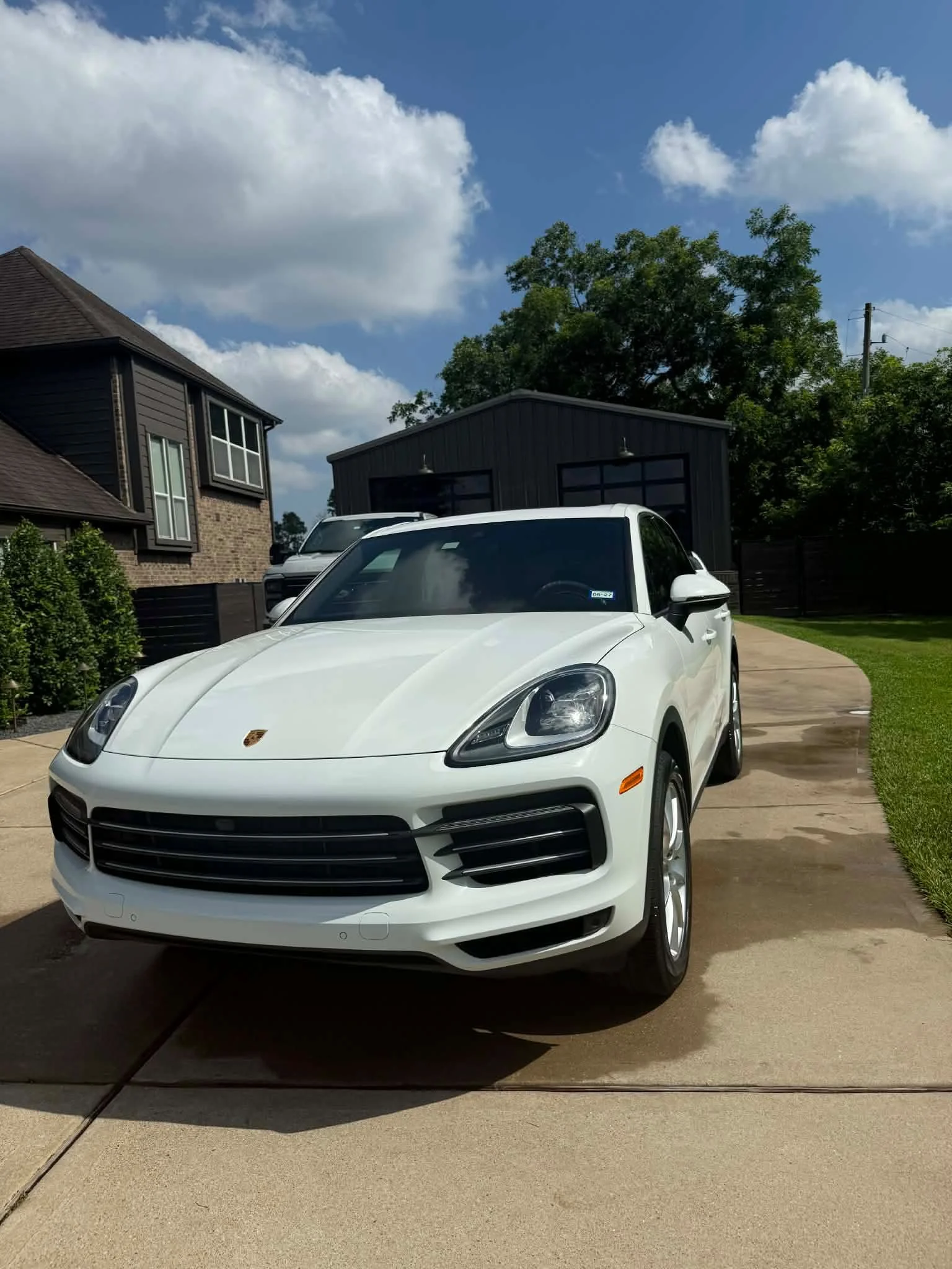White SUV car parked on a driveway in front of a house with a black building and trees in the background under a blue sky with scattered clouds.