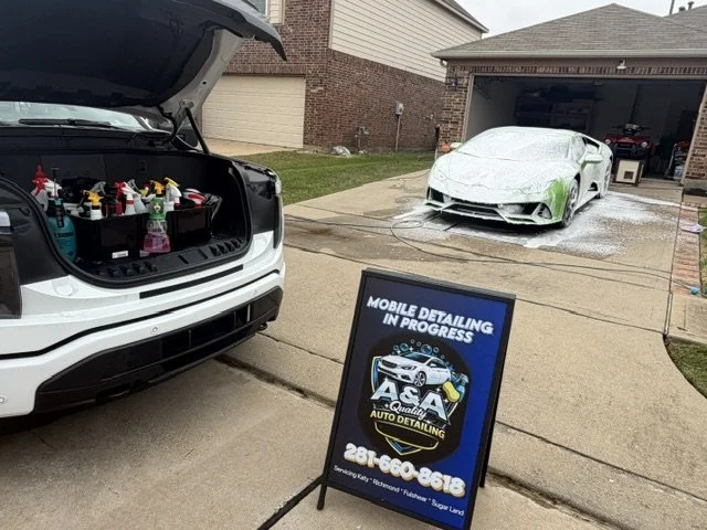 A white SUV with its hood open showing cleaning supplies inside, parked in front of a house. Next to it, a sign advertising mobile auto detailing services. On the driveway, a car is being washed and foamed, with soap and water on the ground.