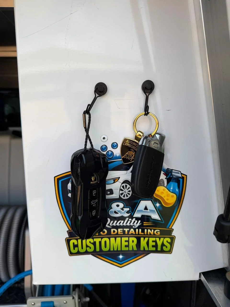 Two sets of car keys hanging on hooks on a white surface with stickers, including a car detailing business logo and a Lamborghini keychain.