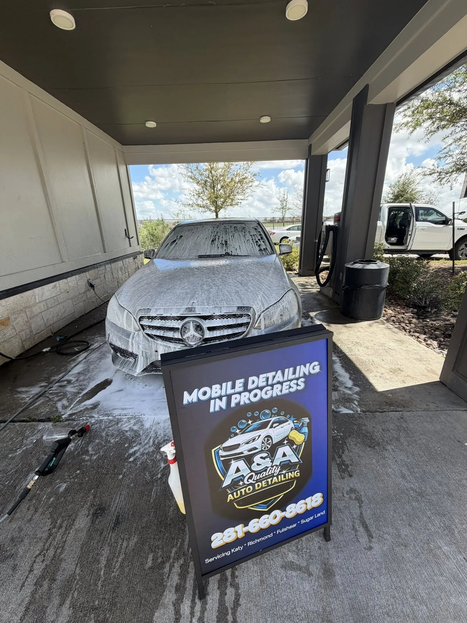 A car covered in soap during an auto detailing process at a mobile service station. There is a signboard in the foreground advertising auto detailing services, with a phone number and locations listed. The station is outdoors with blue skies, clouds, trees, and other vehicles in the background.