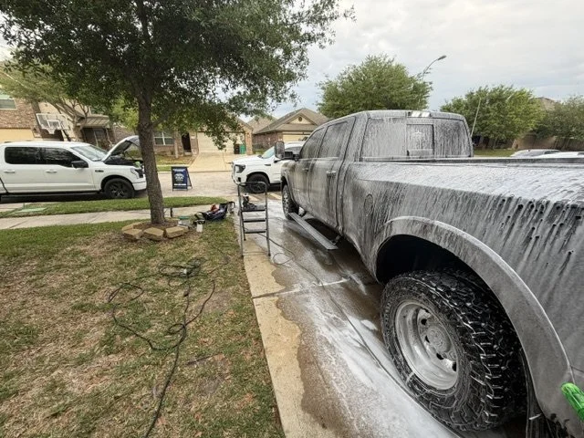 A large pickup truck covered in soap suds during a car wash in a driveway, with other vehicles and houses visible in the background.