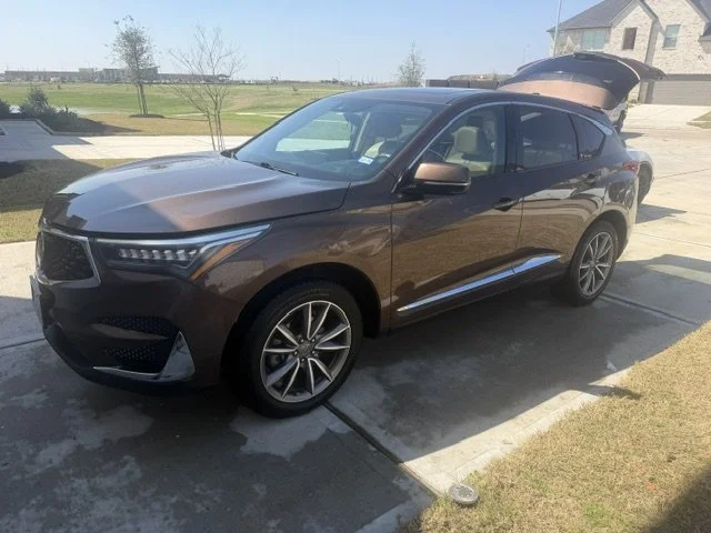 Brown SUV parked on driveway with trunk open, in a suburban yard with grass, trees, and a house nearby.