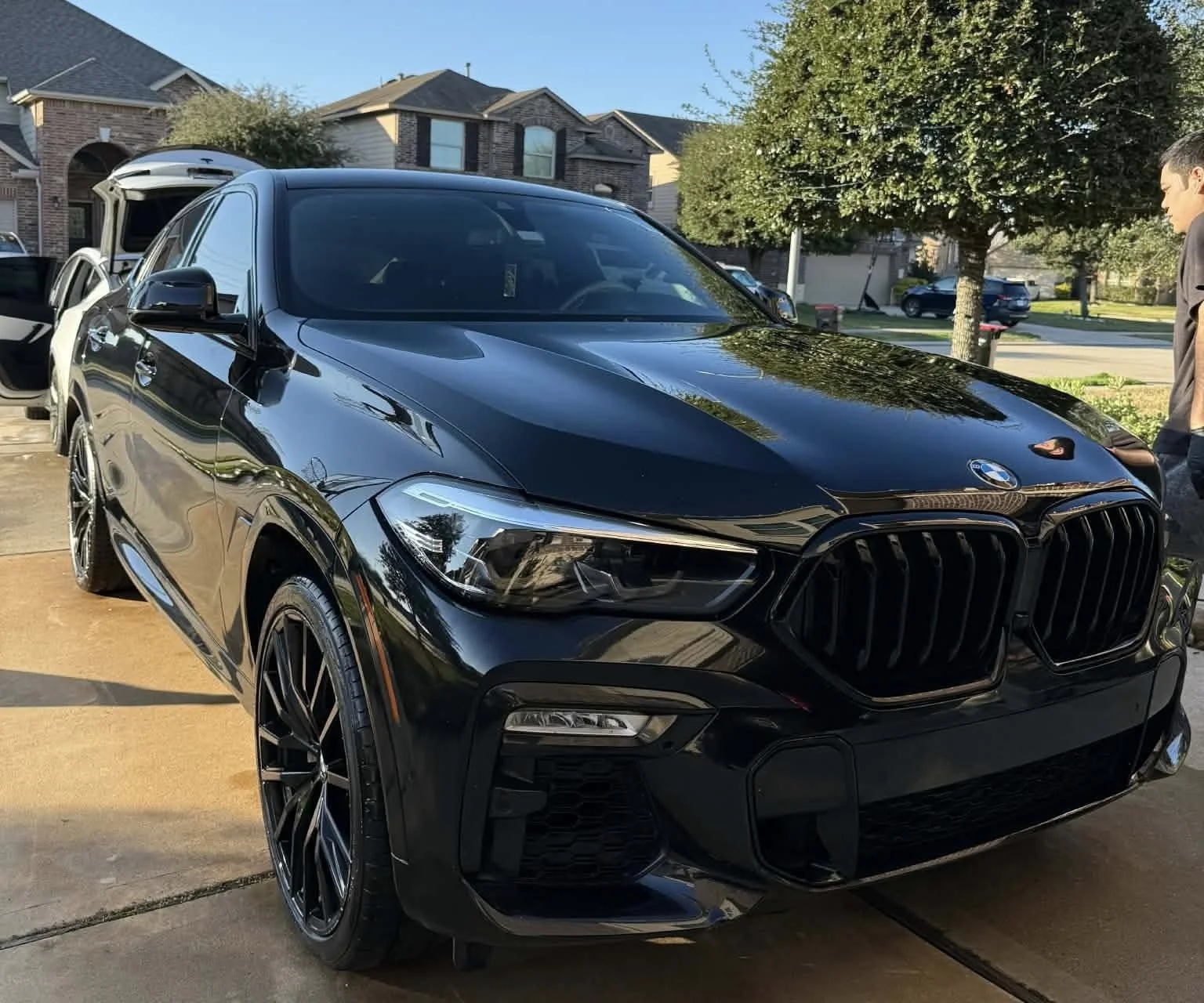 A shiny black BMW SUV parked in a driveway with the driver's side door open, house and trees in the background, and a man standing nearby.
