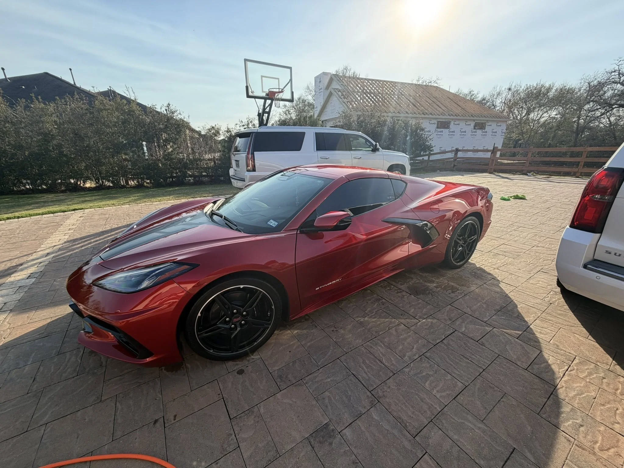 Red sports car parked on a brick driveway next to a white SUV with a basketball hoop attached to its roof. A house under construction and trees in the background.