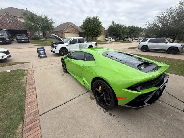 Bright green sports car parked on driveway at car show with various vehicles and houses in the background.