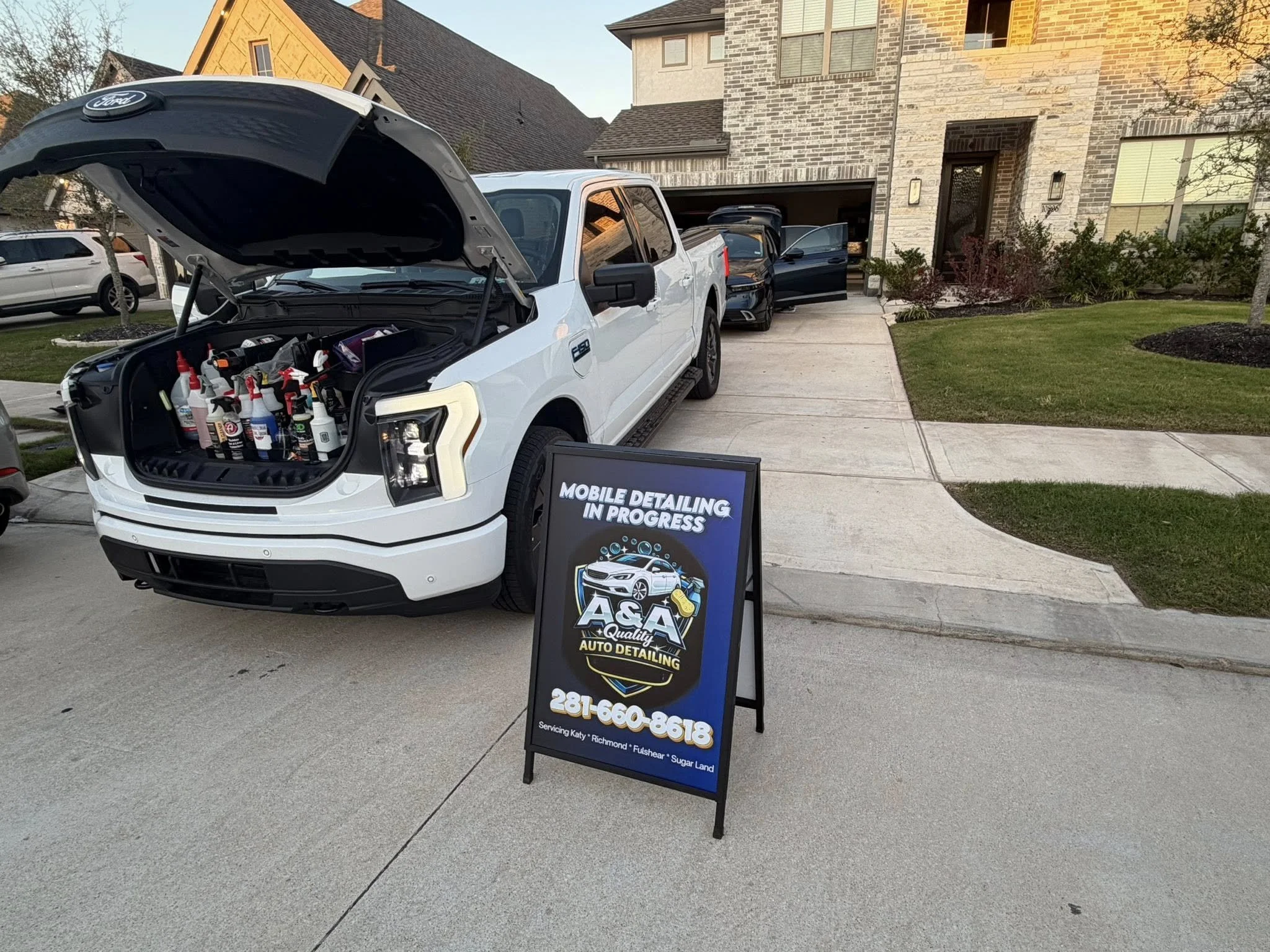 A white pickup truck with its hood open parked in front of a house. There is a signboard advertising auto detailing services and a car in the driveway.