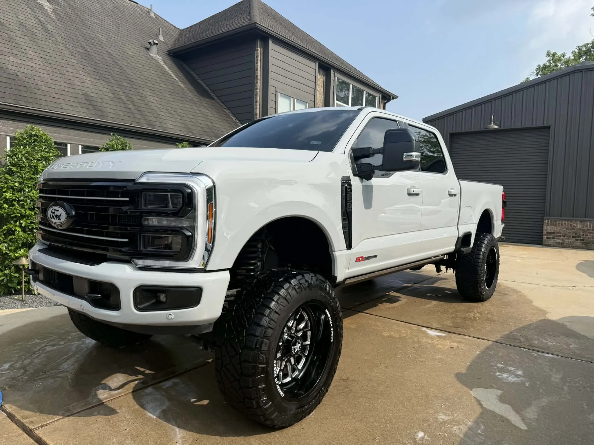 A white Ford Super Duty pickup truck with large black off-road tires is parked in a driveway in front of a modern house with dark gray siding and a gray garage door. The house has a steep roof with multiple angles and small outdoor lights. The sky is partly cloudy.