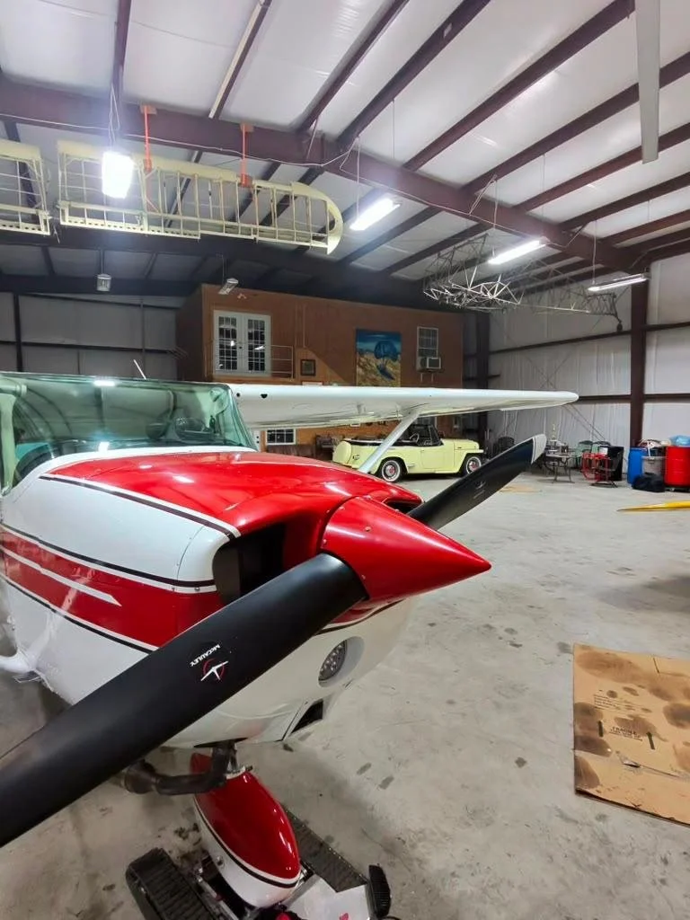 Interior of an aircraft hangar with a small red and white airplane in the foreground, and a vintage yellow car and a second aircraft in the background.