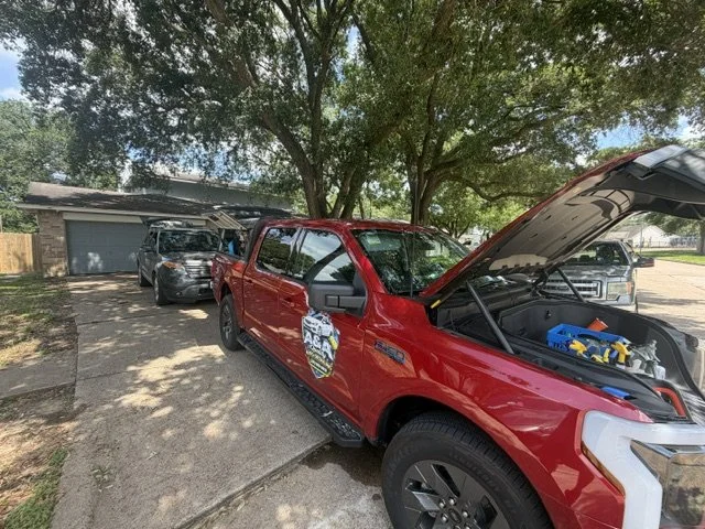 A red police SUV with its hood open, parked on a driveway under a large tree, with other vehicles and a garage in the background.