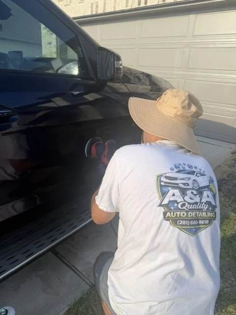 A person wearing a wide-brimmed hat and a white T-shirt with an auto detailing company logo is polishing the side of a black car with a handheld buffer or polishing tool.