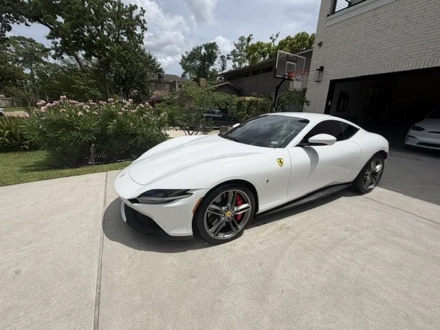 White Ferrari sports car parked on a driveway outside a residential house with a basketball hoop, green trees, and bushes nearby.