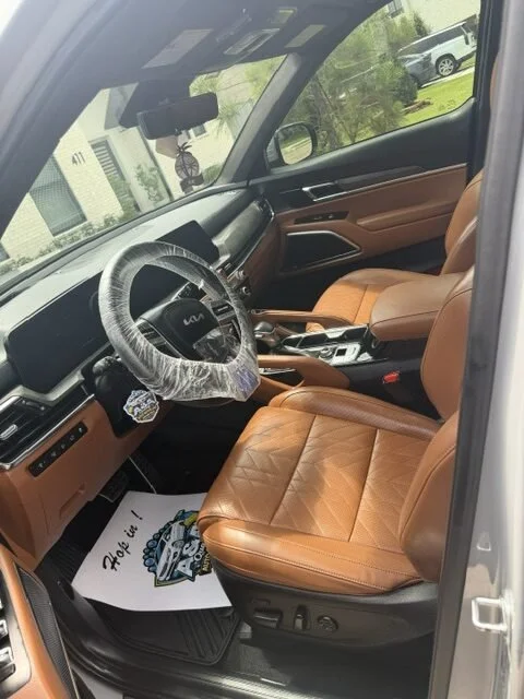 Interior of a vehicle with tan leather seats, a steering wheel wrapped in plastic, and a gear shift. There is a floor mat with a car graphic and the phrase "Thanks for choosing us!" visible on the driver's side.