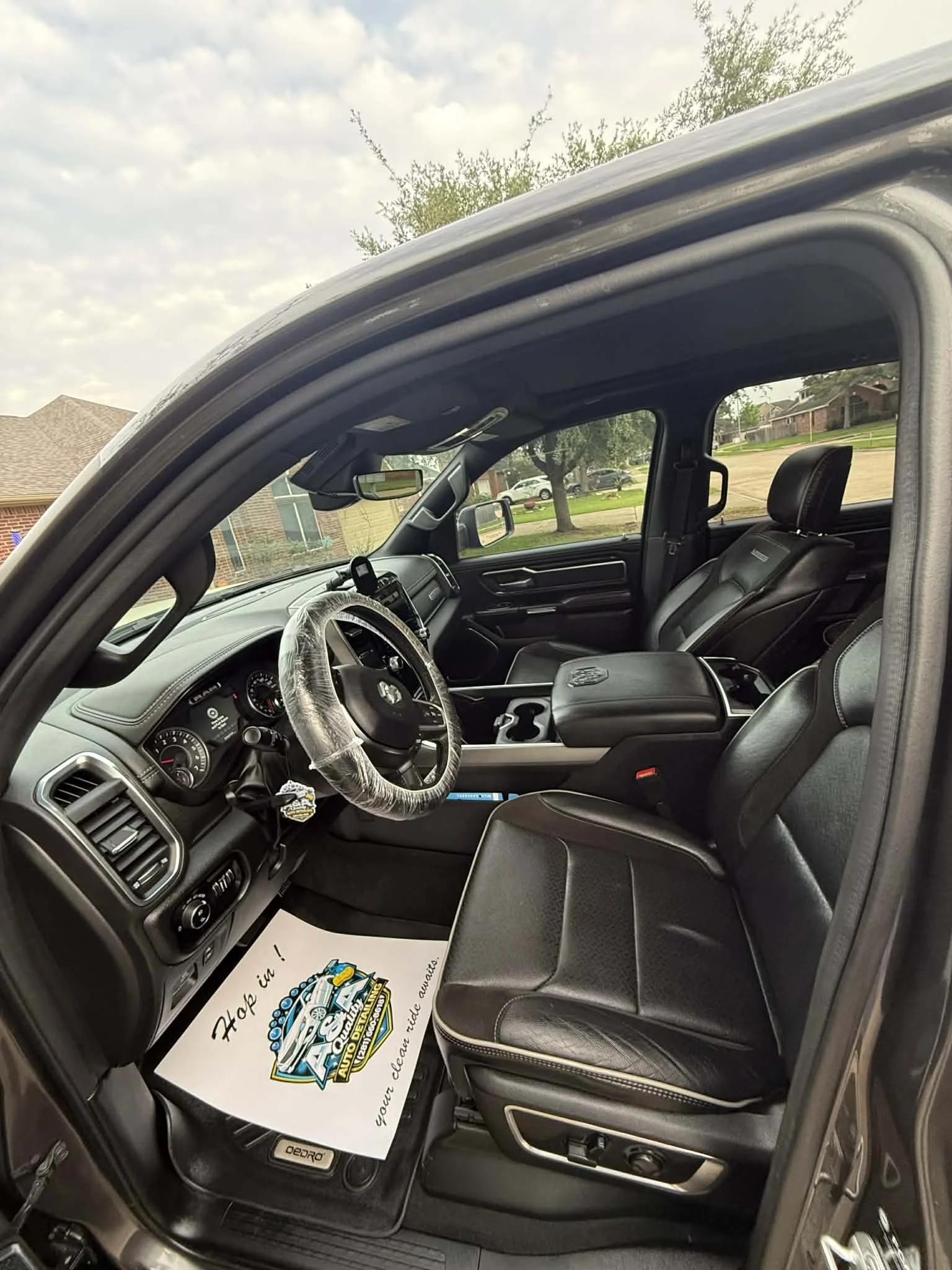 Inside a vehicle showing black seats, dashboard, and steering wheel with a plastic cover, view outside through windows with houses and trees, and a floor mat with a colorful logo and text.