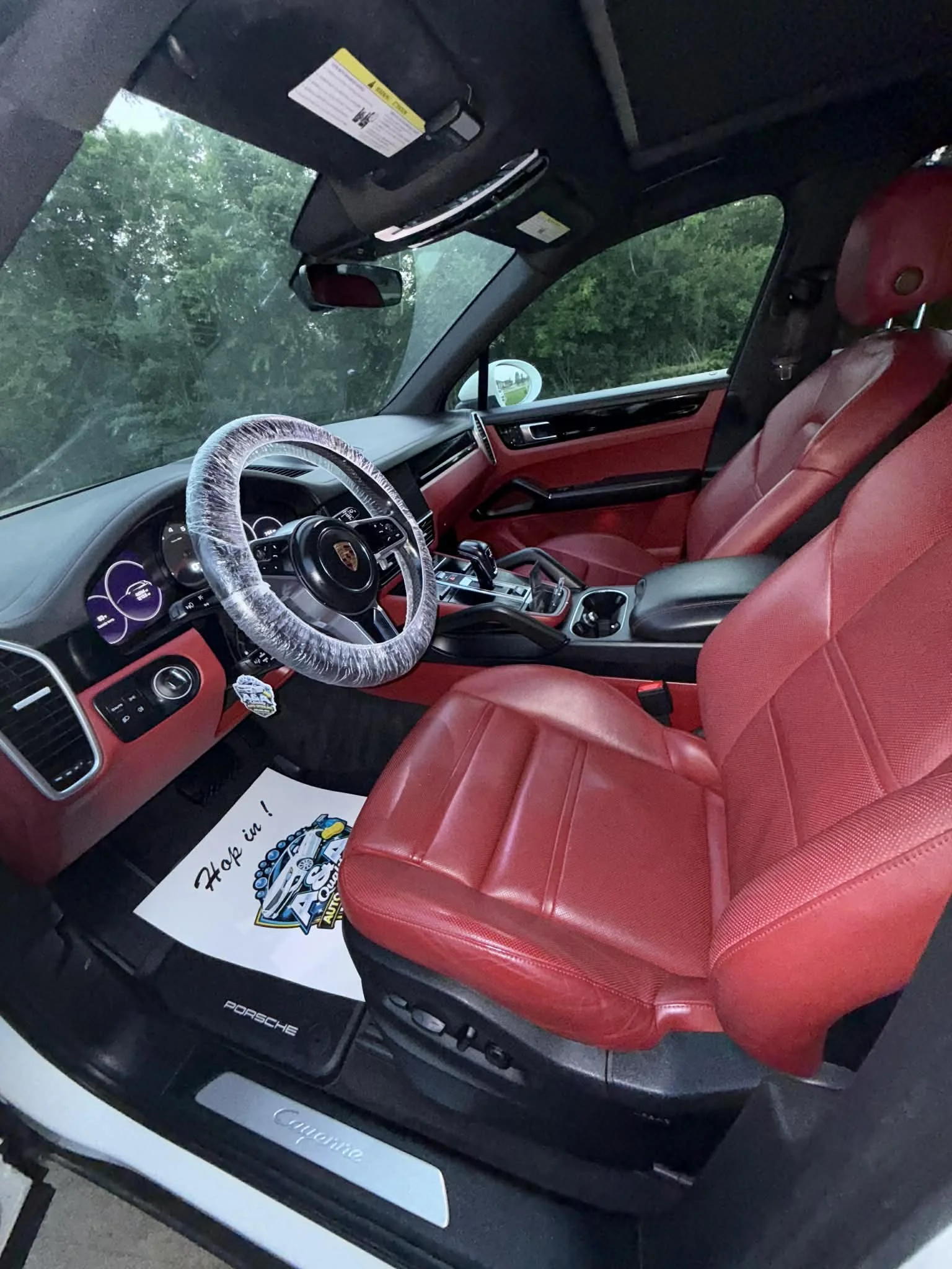 The interior of a Porsche vehicle featuring red leather seats, a white plastic-wrapped steering wheel with the Porsche emblem, a digital display, and a black dashboard with various controls. The black floor mat has a Porsche logo and a white paper mat with a cartoon character and the text 'Hooray!'