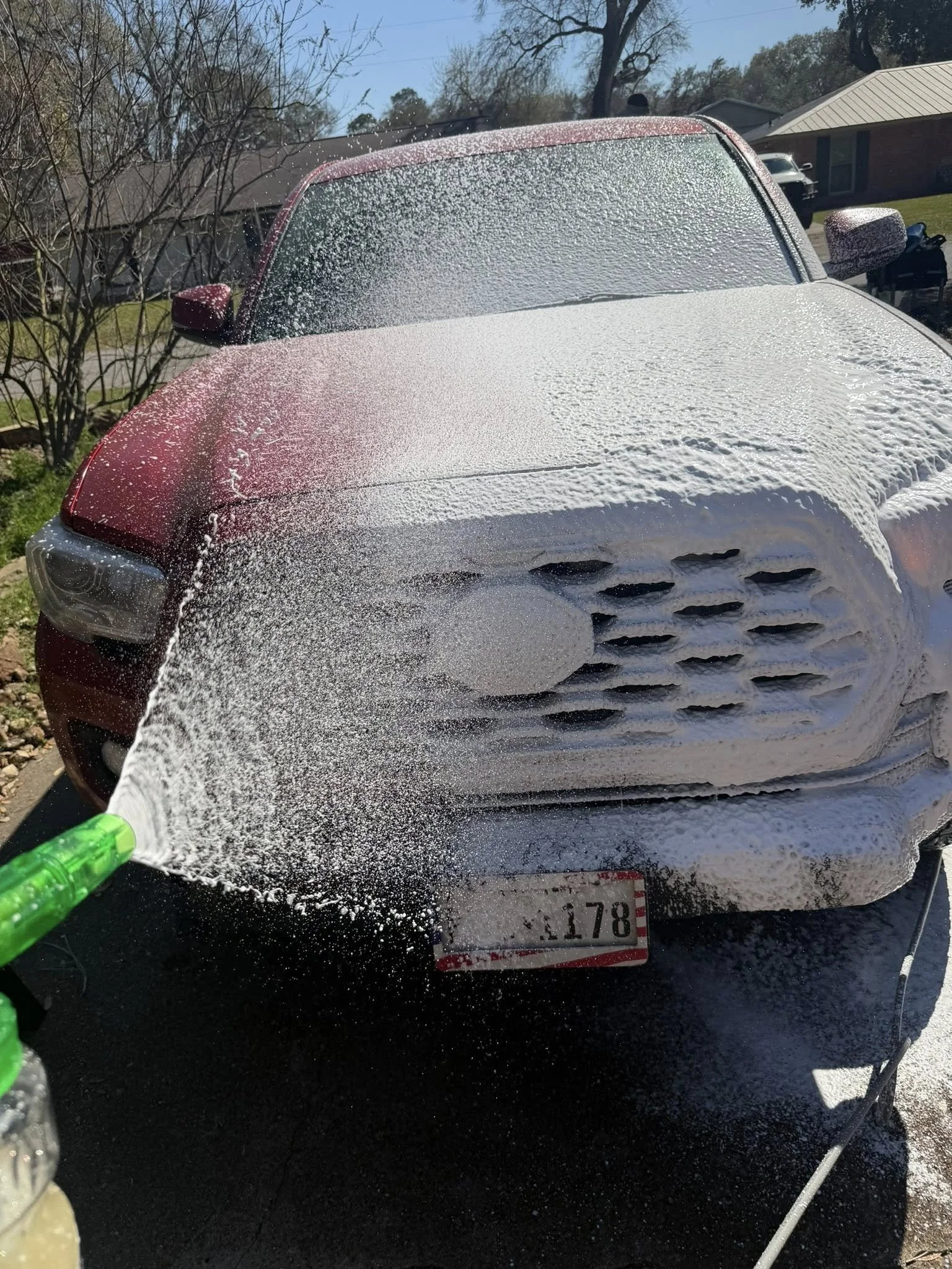 Front of a red car being washed with soap and water, with a garden and houses in the background.