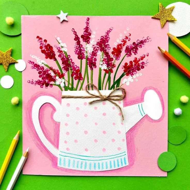 Colorful craft project featuring a pink card with a hand-painted watering can holding pink and white flowers, surrounding decorative elements like stars and circles.