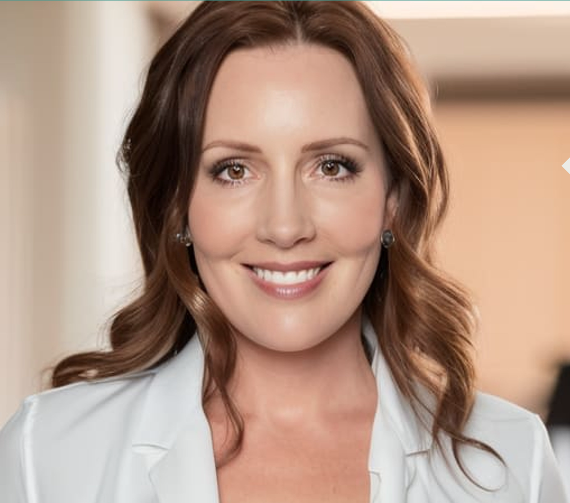 A woman with medium-length wavy brown hair and light brown eyes, smiling, wearing earrings and a white blouse.